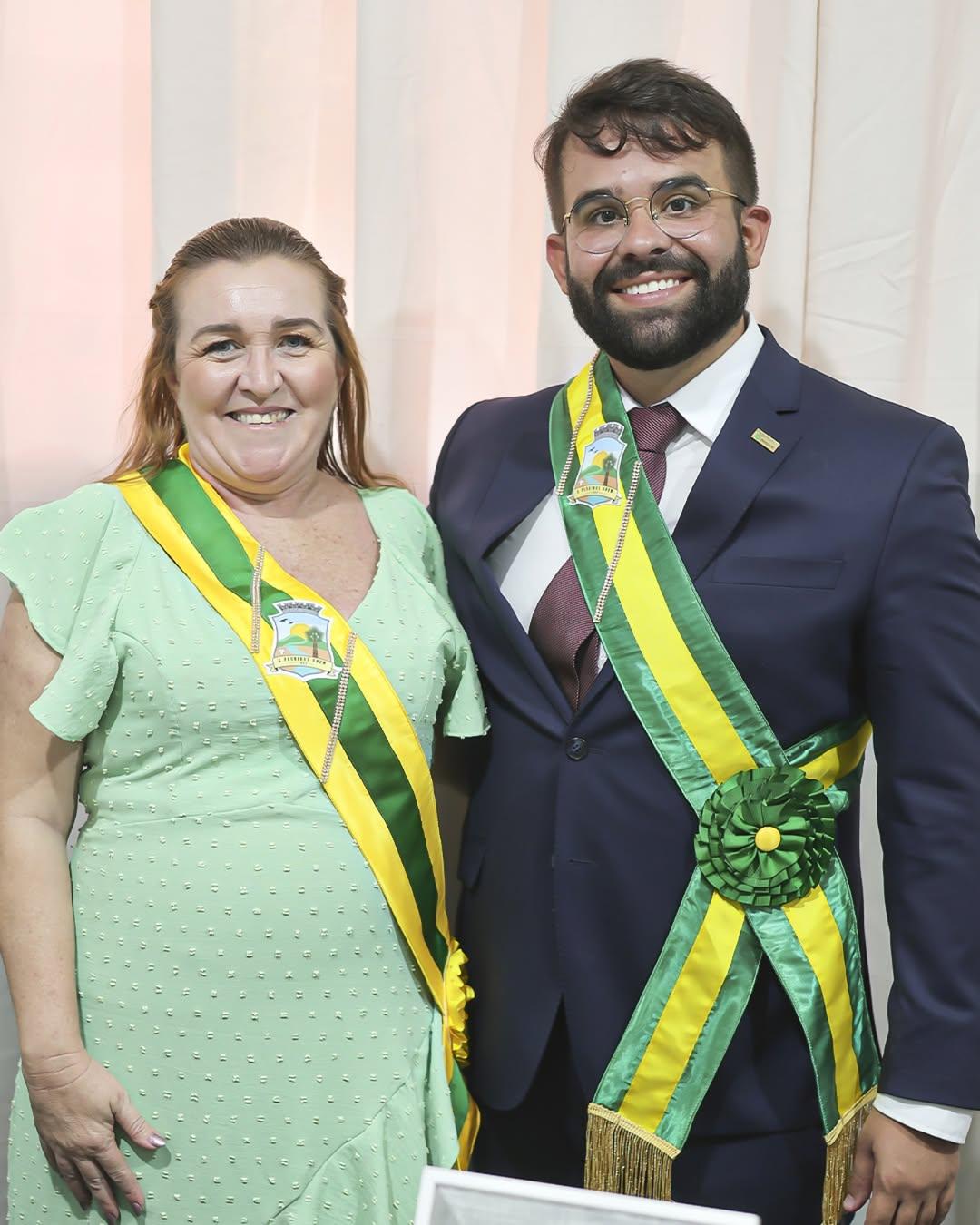 Mulher e homem posam sorridentes lado a lado em frente a uma cortina clara. Ambos usam faixas cerimoniais nas cores verde, amarelo e branco com o brasão do município de Moraújo-CE. A mulher veste um vestido verde claro com bolinhas e a faixa atravessa seu ombro direito. O homem usa terno azul-marinho com gravata vinho, óculos e barba, e a faixa atravessa seu ombro esquerdo, com uma roseta verde à cintura. Ambos aparentam participar de uma cerimônia oficial.