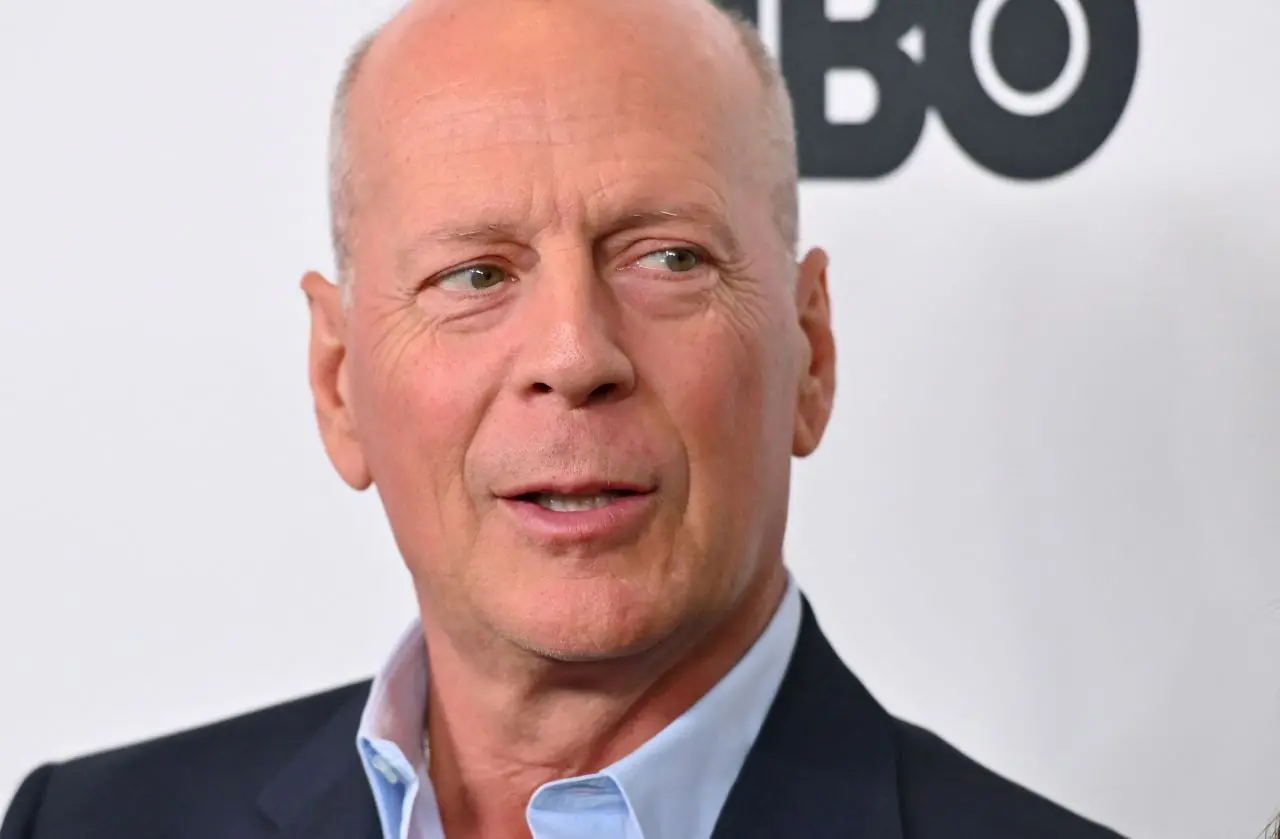Bruce Willis sorrindo, diante de fundo branco. ele está olhando para o lado direito.