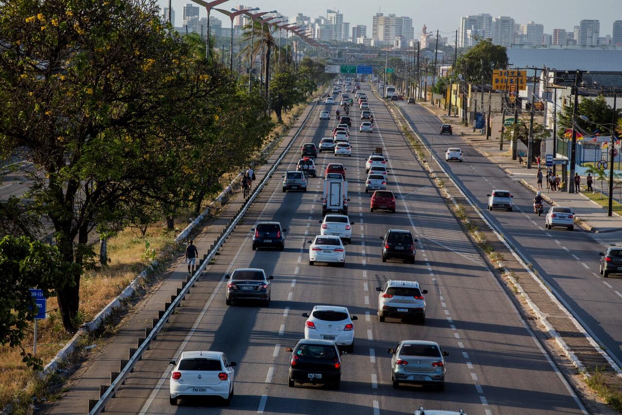 Foto mostra trecho urbano da BR-116 em Fortaleza, com tráfego intenso de veículos e ciclovia à esquerda. Ao fundo, vê-se o horizonte com edifícios altos e vegetação nas laterais da via.