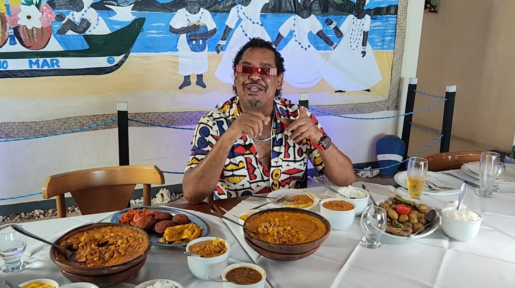 Compadre Washington, cantor do grupo É o Tchan, aparece sorridente sentado à mesa em um restaurante decorado com temática afro-brasileira. Ele veste uma camisa estampada colorida e óculos vermelhos estilosos. À sua frente, uma mesa farta com pratos típicos da culinária baiana, como vatapá, acarajé, caruru, arroz e moquecas em panelas de barro. Ao fundo, um mural pintado retrata figuras negras em trajes brancos religiosos, remetendo ao candomblé, com o mar azul ao fundo. O clima é festivo e cultural.