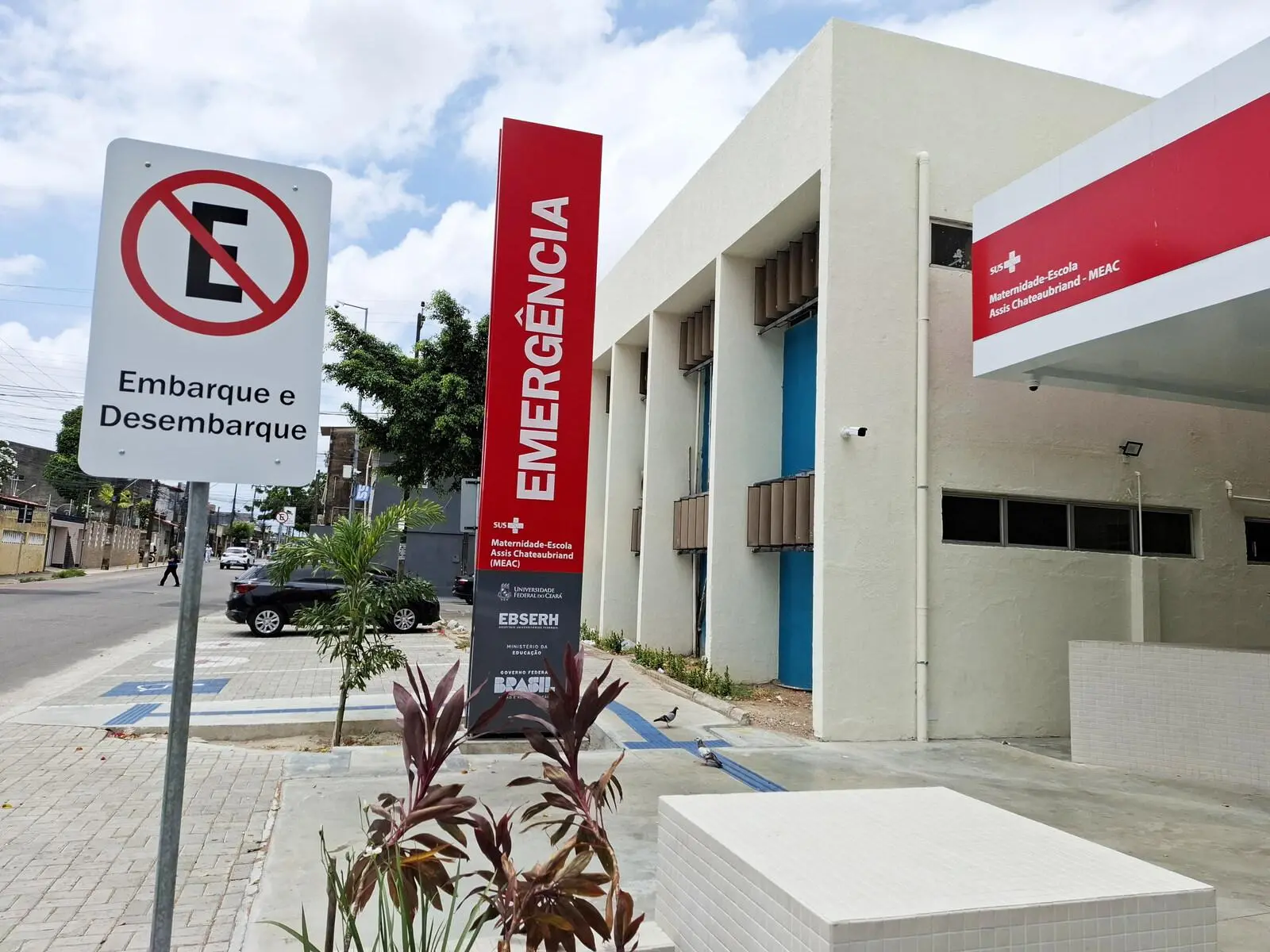 A imagem mostra a fachada da Maternidade Escola Assis Chateaubriand, com uma placa alta e vermelha indicando EMERGÊNCIA. Há também uma placa de sinalização de trânsito de Proibido Estacionar com a indicação Embarque e Desembarque. Um carro preto e algumas pessoas são visíveis na rua ao lado.