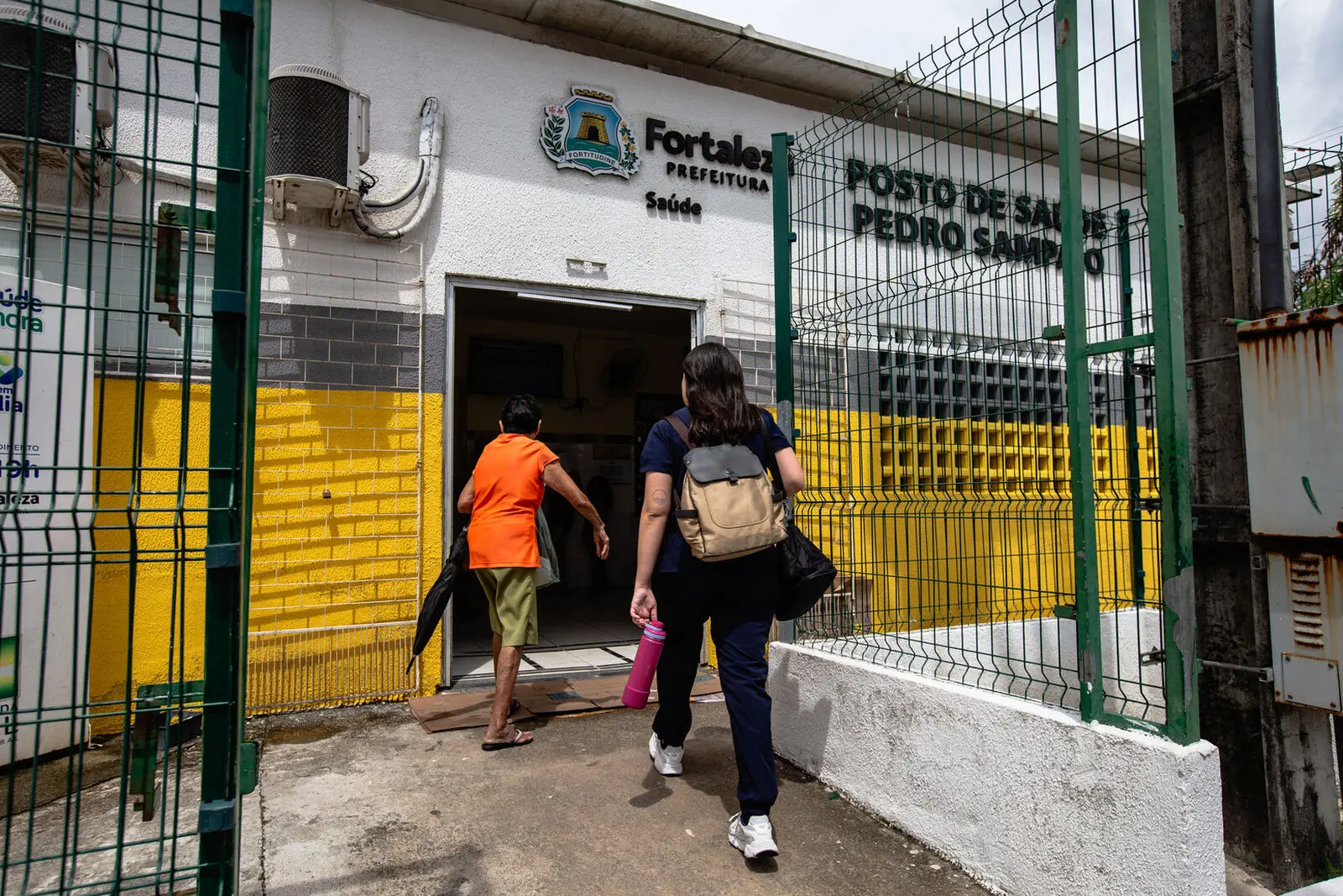 Imagem mostra fachada de um posto de saúde de Fortaleza. Um homem e uma mulher estão de costas entrando na unidade.
