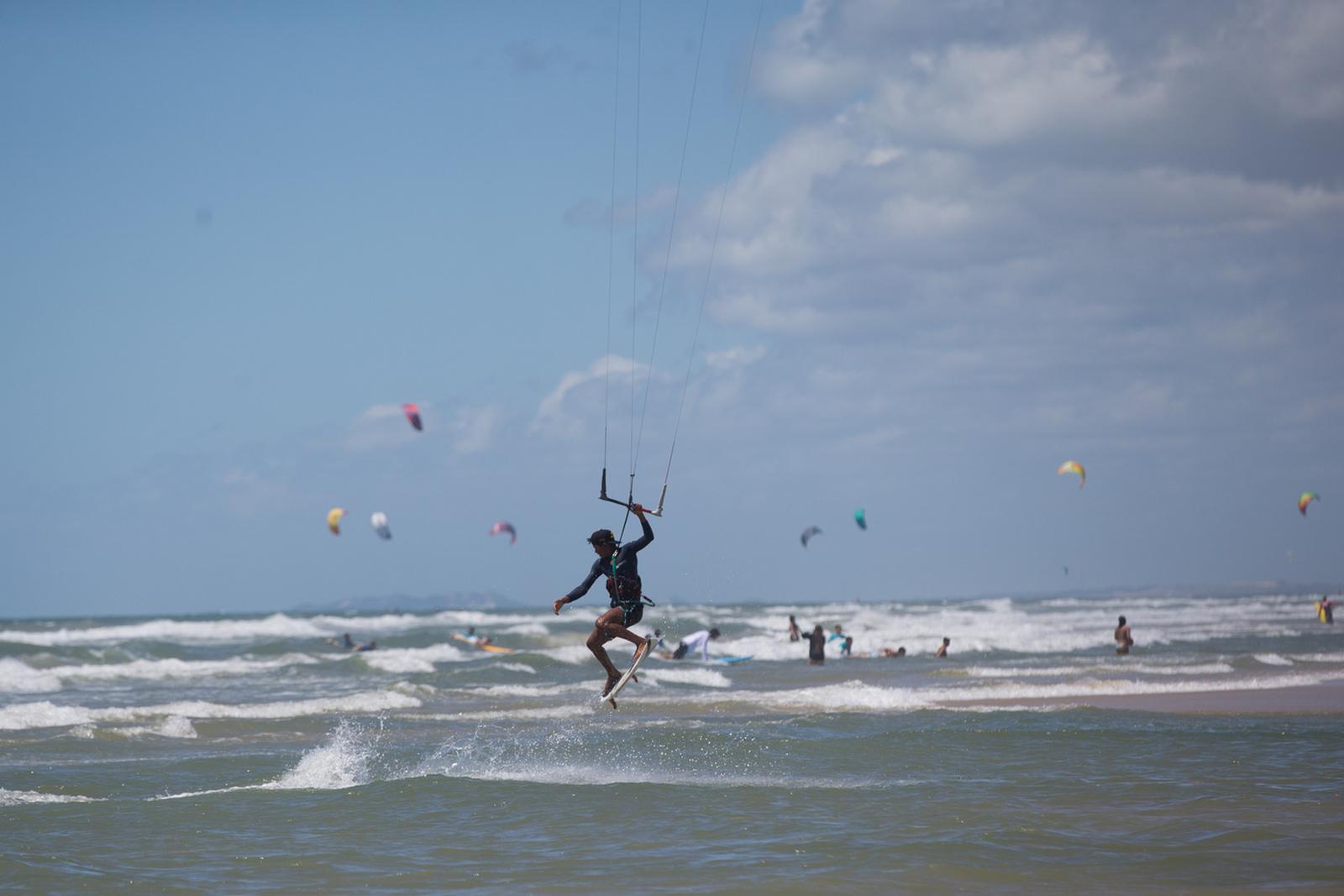 A imagem mostra uma pessoa praticando kitesurf, elevando-se sobre a água do mar. O céu é azul com nuvens esparsas, e várias pipas coloridas de kitesurf são visíveis à distância sobre o oceano. Ondas quebram na praia, onde outras pessoas estão na água e na areia, no Ceará.