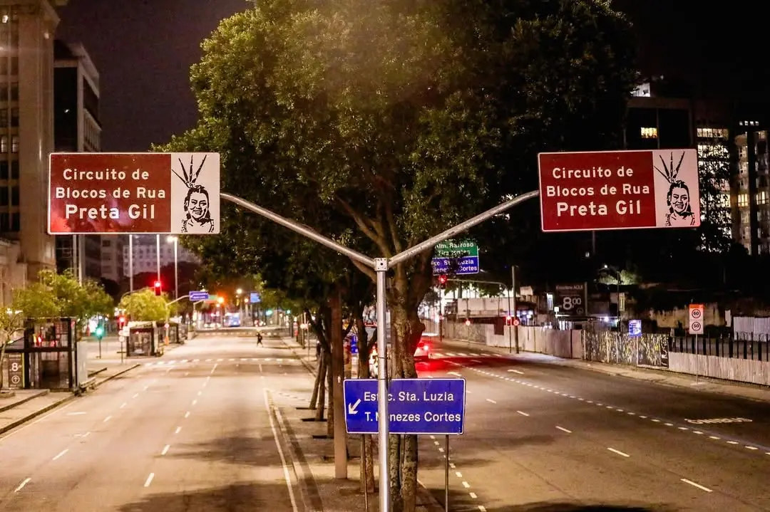 Imagem de rua vazia à noite com placas de trânsito, incluindo uma com grafite de mulher com cabeça de espeto, indicando o Circuito de Blocos de Rua Preta Gil no Rio de Janeiro