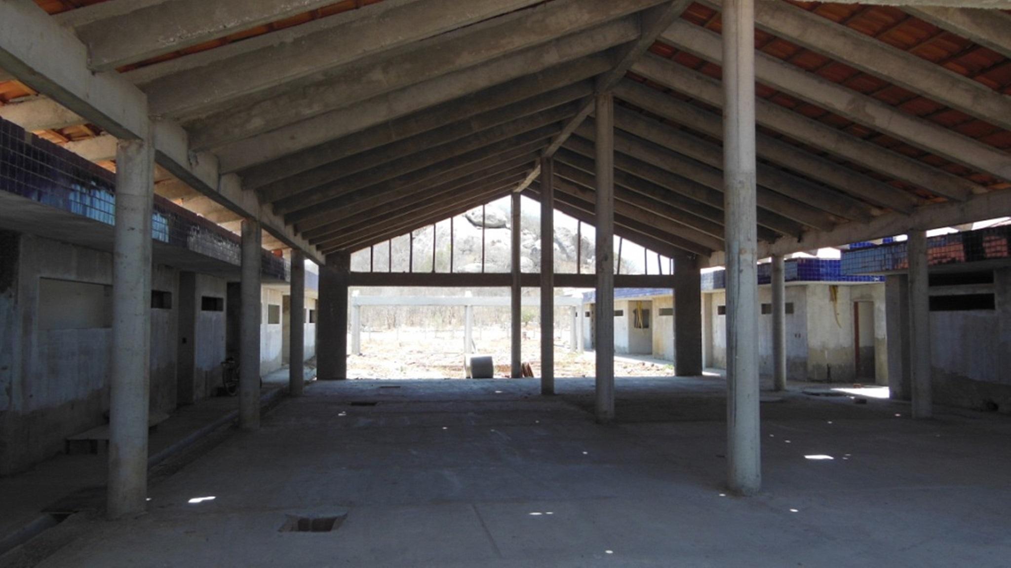 Foto mostra o interior de um prédio em construção, com estrutura de concreto, telhado instalado e piso ainda bruto. O local aparenta ser uma escola com salas distribuídas nas laterais.