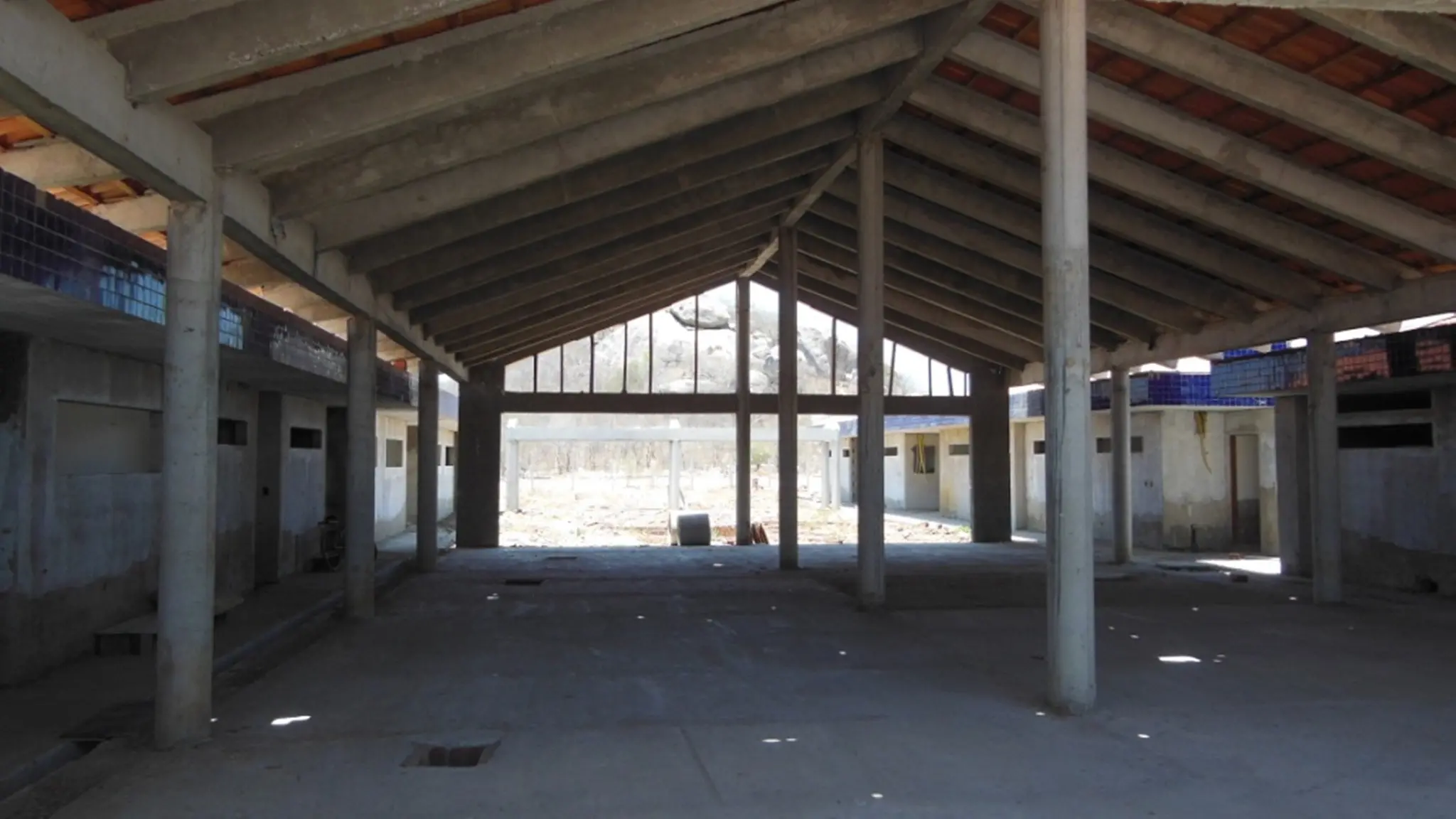 Foto mostra o interior de um prédio em construção, com estrutura de concreto, telhado instalado e piso ainda bruto. O local aparenta ser uma escola com salas distribuídas nas laterais.