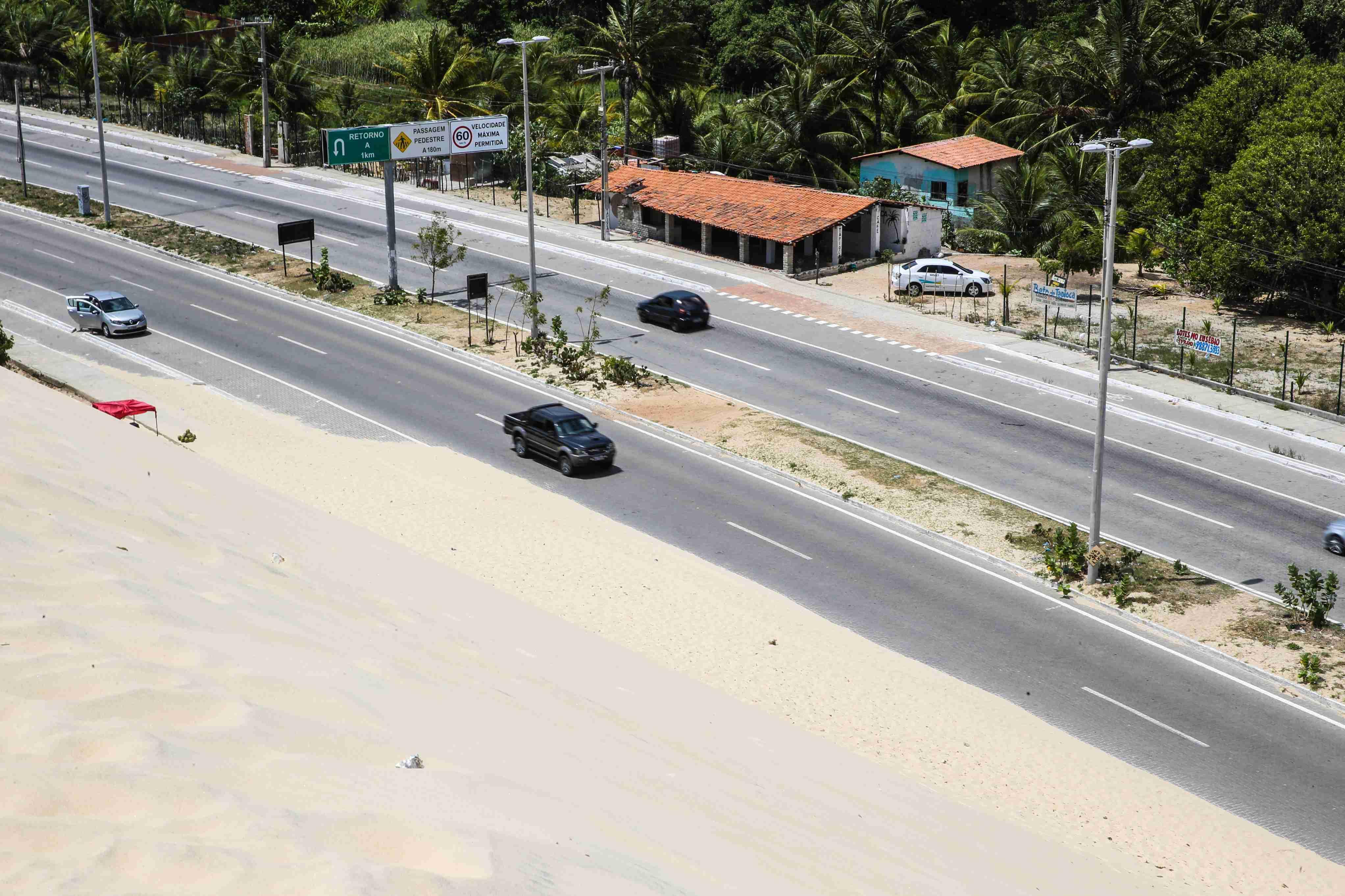 Uma visão aérea de uma rodovia asfaltada de múltiplas pistas, dividida por dunas de areia, sob um céu claro. Carros brancos e pretos trafegam na rodovia, que tem postes de luz ao longo de seu comprimento. Há casas com telhados de telha e vegetação densa visíveis ao lado da estrada.
