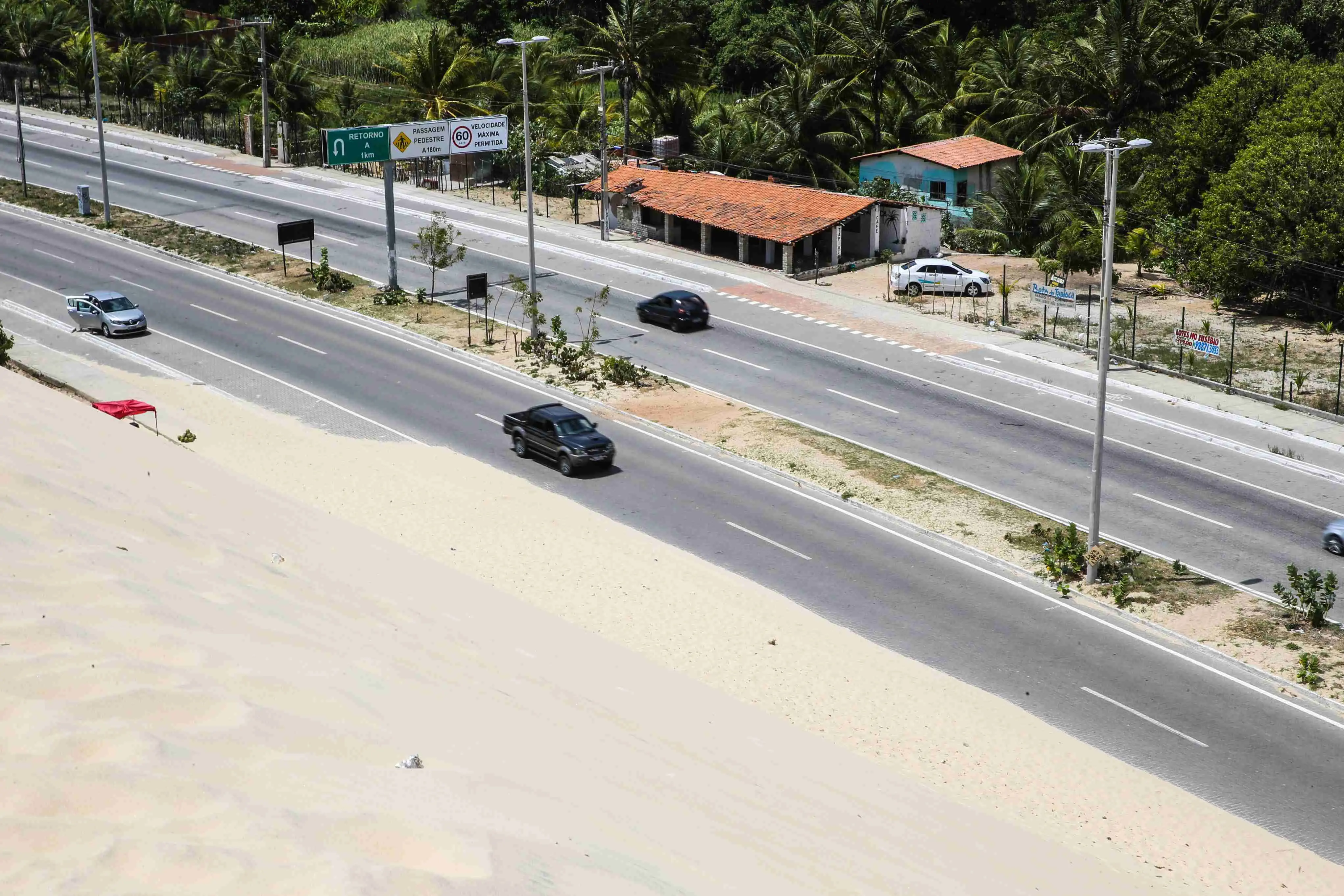 Uma visão aérea de uma rodovia asfaltada de múltiplas pistas, dividida por dunas de areia, sob um céu claro. Carros brancos e pretos trafegam na rodovia, que tem postes de luz ao longo de seu comprimento. Há casas com telhados de telha e vegetação densa visíveis ao lado da estrada.