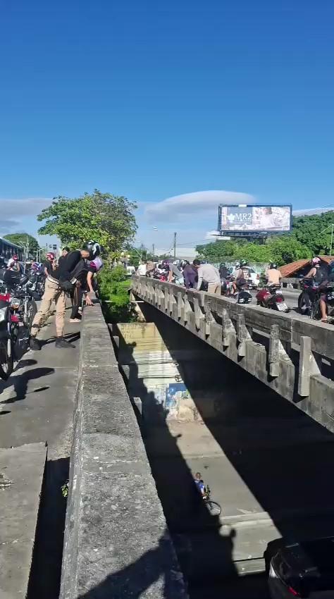 Imagem de um trânsito intenso com veículos e motocicletas parados em uma via urbana, sob céu azul com algumas nuvens e árvores ao redor, indicando congestionamento.