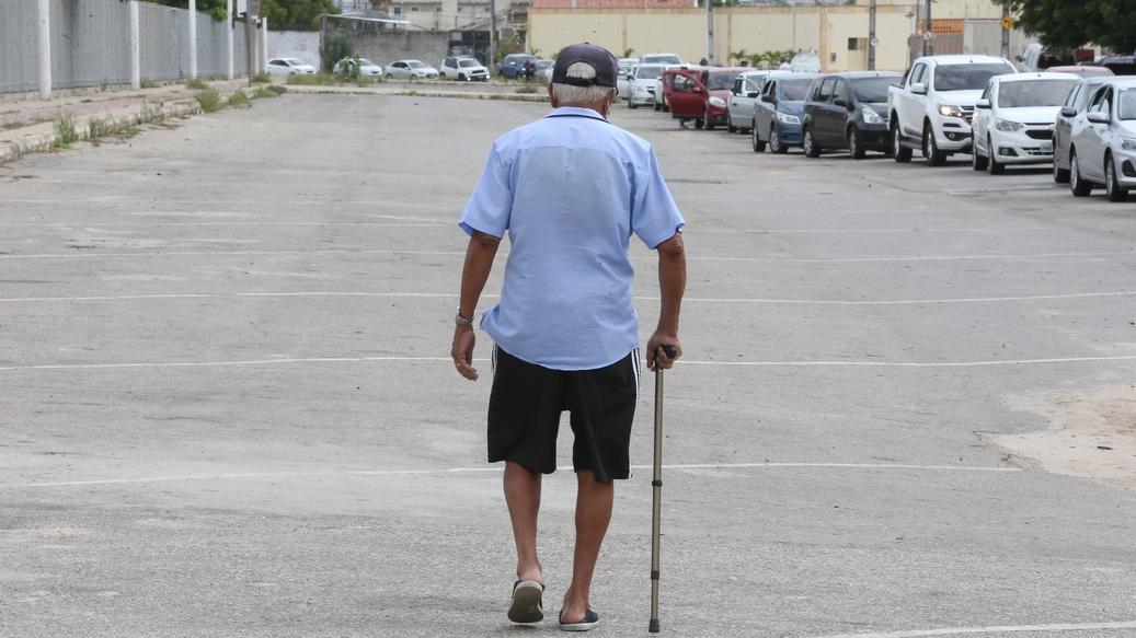 Homem idoso visto de costas caminhando em um estacionamento vazio, usando bengala na mão direita. Ele veste camisa azul clara, bermuda preta, boné escuro e chinelos. À direita da imagem, há uma fila de carros estacionados.
