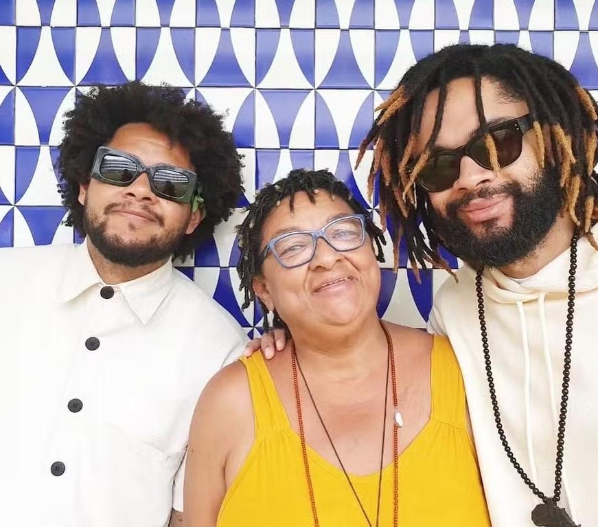 Imagem da Dona Jacira, mãe de Emicida e Evandro Fióti, que morreu aos 60 anos. Ela está entre os dois filhos, é uma mulher negra e vesta camiseta amarela