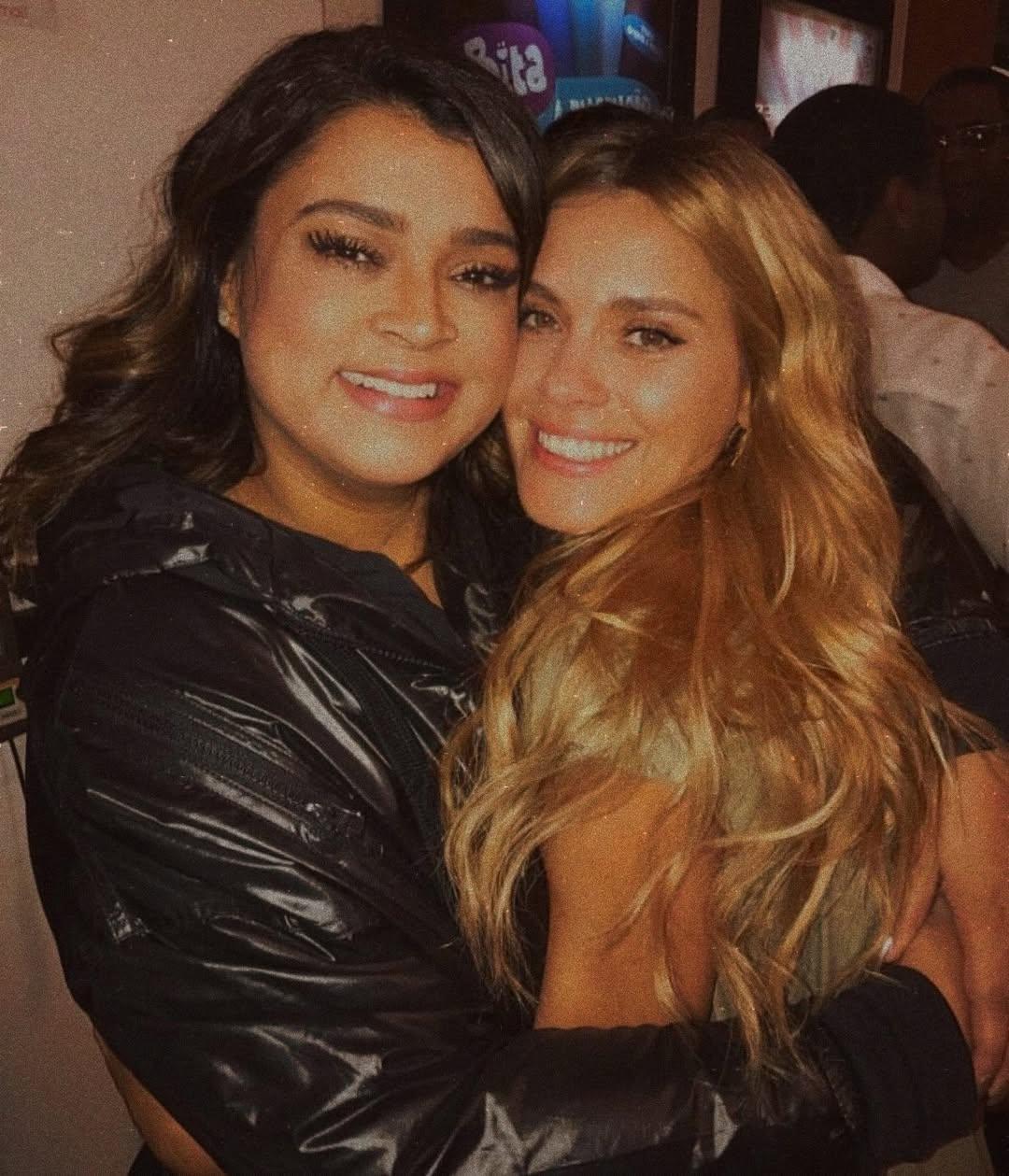 Duas mulheres sorrindo e abraçadas, comemorando juntas em um ambiente animado, com uma atmosfera de festa ou reunião social.