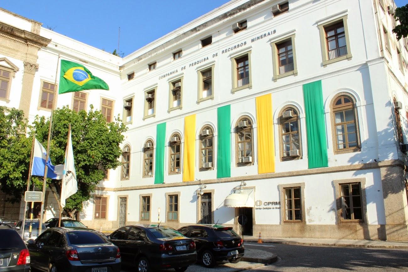 Fachada do prédio da Companhia de Pesquisa de Recursos Minerais (CPRM) no Brasil, decorada com panos nas cores da bandeira nacional, sob céu azul.