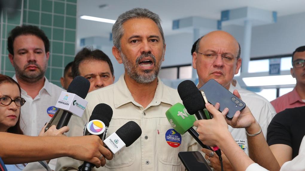A imagem mostra Elmano de Freitas, homem com barba e cabelo grisalhos, usando uma camisa clara, falando para vários microfones de imprensa. Ele está cercado por outras pessoas, algumas das quais seguram celulares.