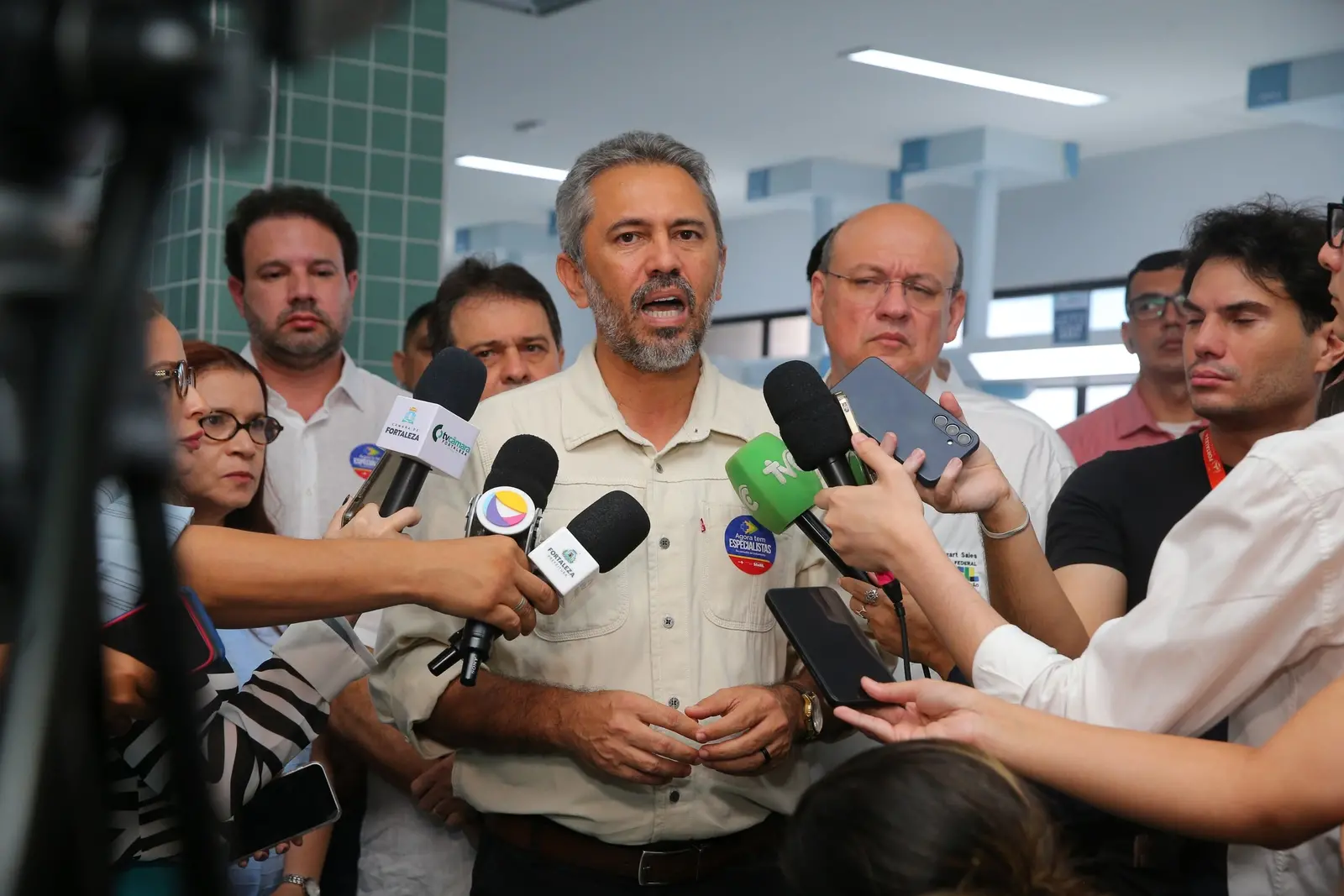 A imagem mostra Elmano de Freitas, homem com barba e cabelo grisalhos, usando uma camisa clara, falando para vários microfones de imprensa. Ele está cercado por outras pessoas, algumas das quais seguram celulares.
