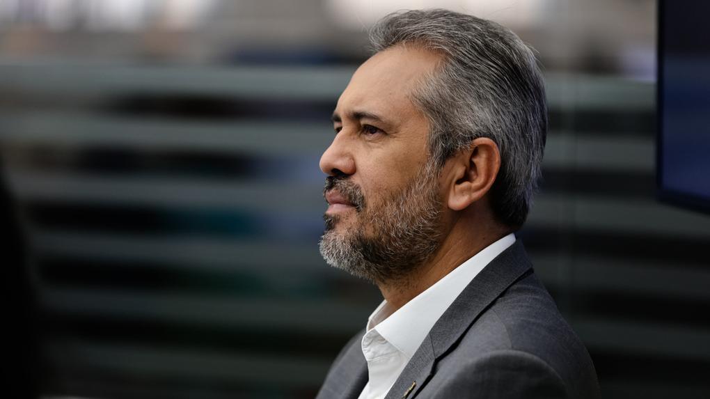 Governador Elmano de Freitas de perfil. Ele é um homem de meia idade, de barba e cabelo grisalhos, e está de terno cinza com camisa social branca