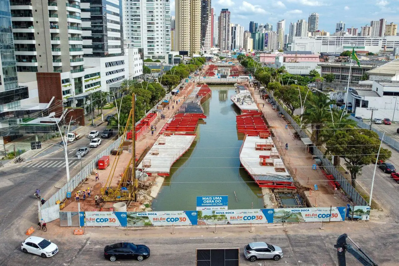 Imagem de um canteiro de obras na cidade, com barracos, máquinas e trabalhadores, ao lado de um canal de água, com arranha-céus ao fundo, em uma área de construção urbana.