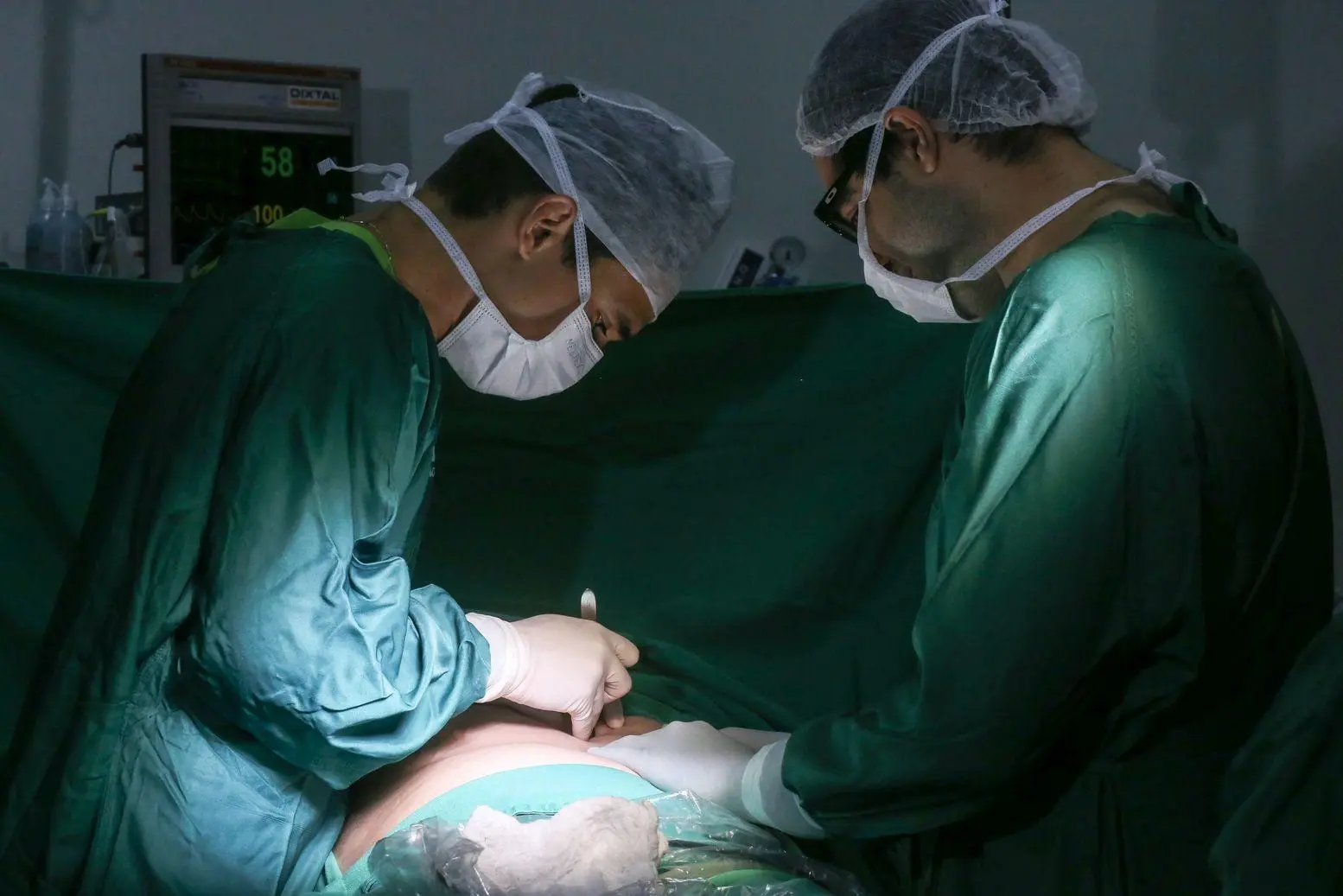 Imagem mostra dois médicos realizando cirurgia em paciente. Eles vestem trajes verdes, touca e máscara hospitalares.