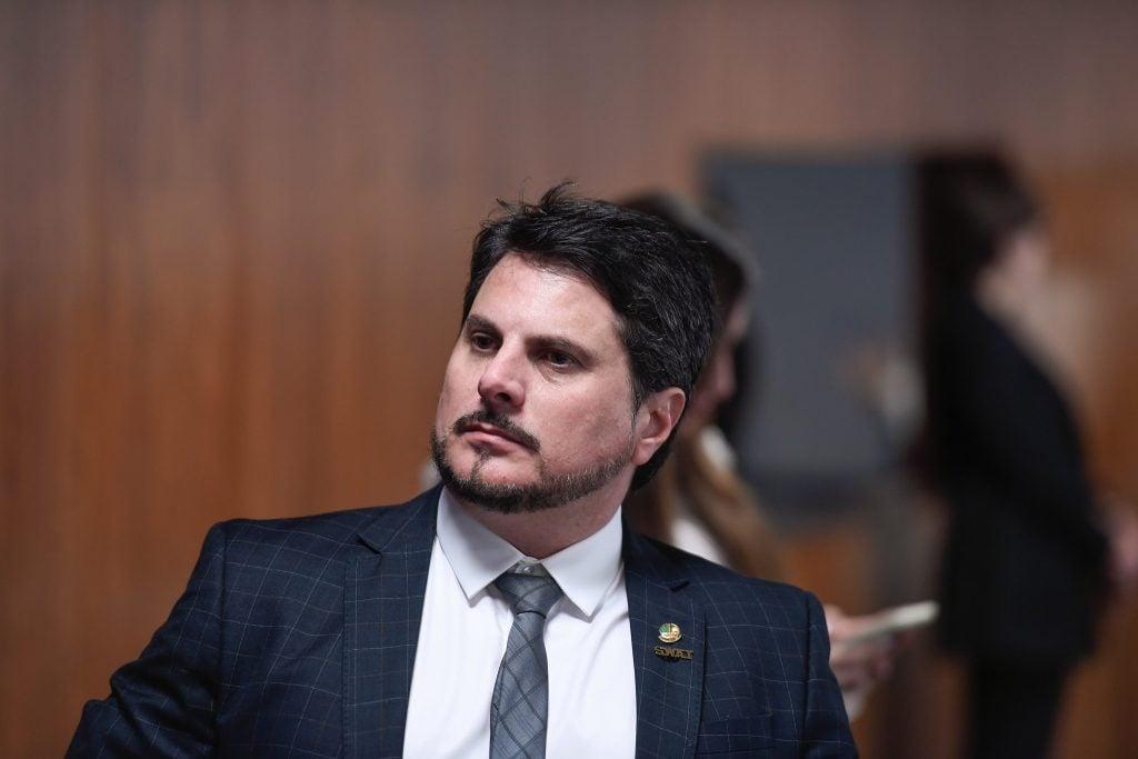 Homem vestido formalmente com terno e gravata em evento social ou reunião, com expressão séria e fundo de ambiente interno com outras pessoas ao fundo.