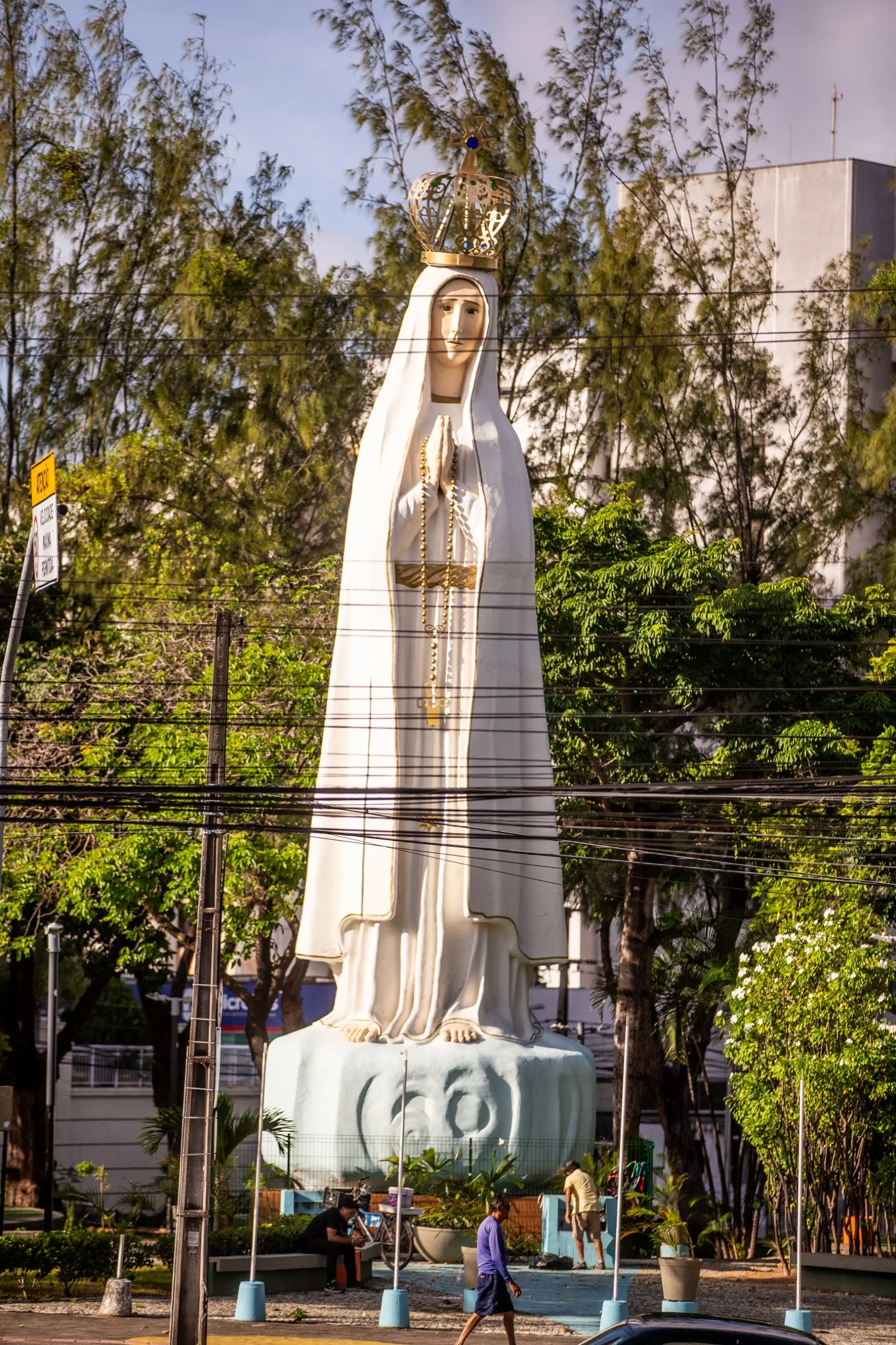 Uma fotografia de uma estátua grande de Nossa Senhora de Fátima em um canteiro central de uma avenida. A estátua é branca com uma coroa dourada e está sobre uma base azul. A imagem é vista por trás de uma rede de fios elétricos e postes. Ao fundo, há muitas árvores e edifícios. Pessoas podem ser vistas no primeiro plano e ao redor da base da estátua.