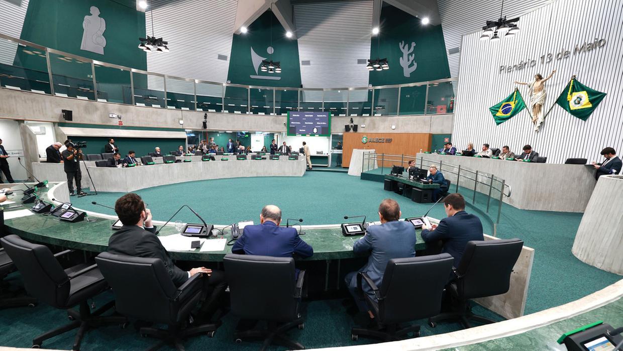 Vista ampla do plenário da Assembleia Legislativa do Estado do Ceará (ALECE) durante uma sessão. A imagem mostra deputados e deputadas sentados em suas cadeiras ao redor de uma grande mesa semicircular, com computadores e microfones à frente. Ao fundo, há uma parede com o nome “Plenário 13 de Maio”, uma escultura de Jesus Cristo com os braços abertos entre as bandeiras do Brasil e do Estado do Ceará. O ambiente é moderno, com tons de verde predominantes, e várias pessoas acompanham a sessão. Uma equipe de imprensa registra o momento.