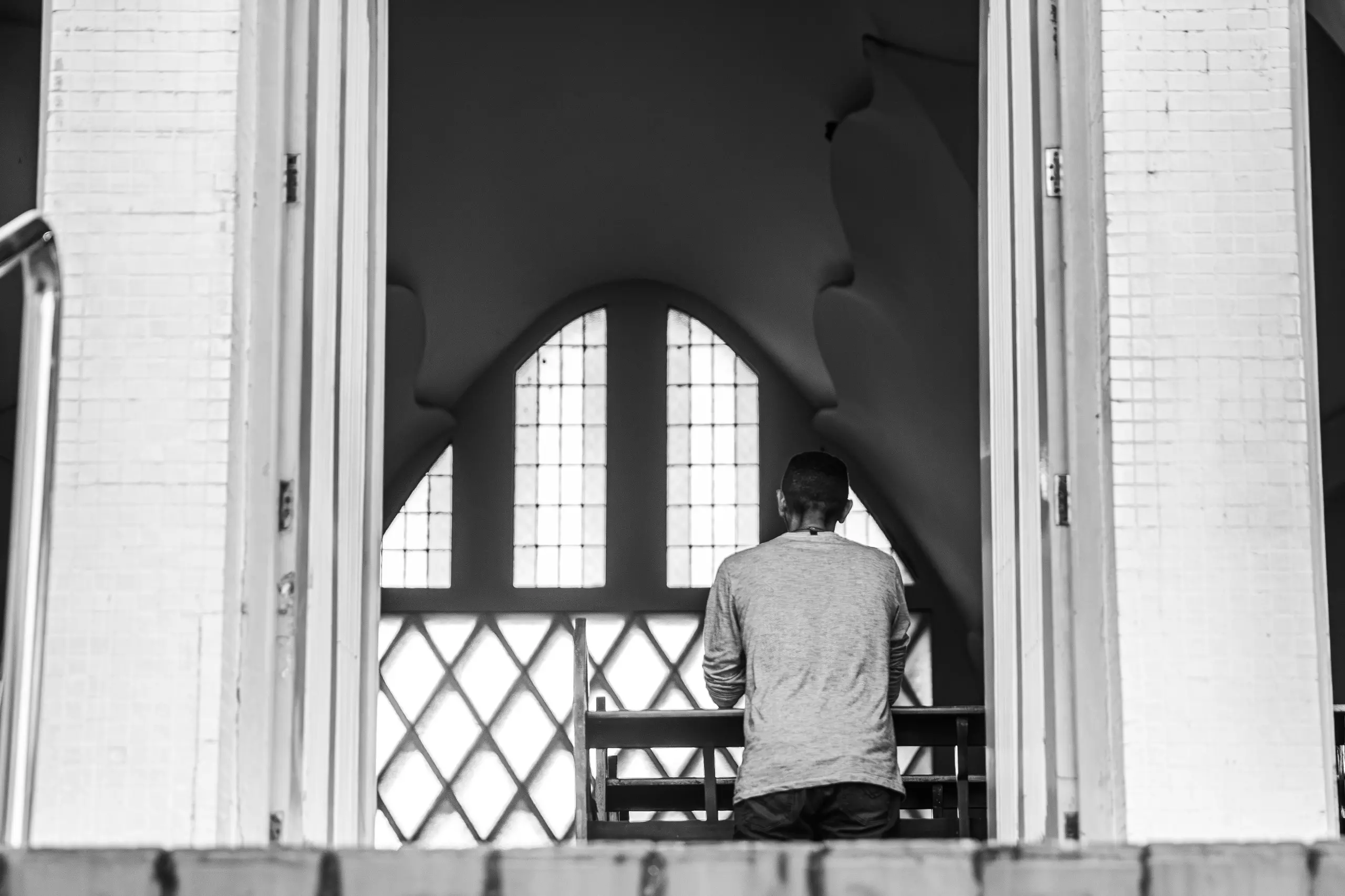 Uma fotografia em preto e branco de um fiel rezando dentro da igreja. A imagem é vista de fora, através de uma porta aberta, mostrando uma pessoa de costas, de joelhos, voltada para uma grande janela em arco com vitrais de vidro. A arquitetura interna da igreja, com suas linhas curvas, é visível ao fundo