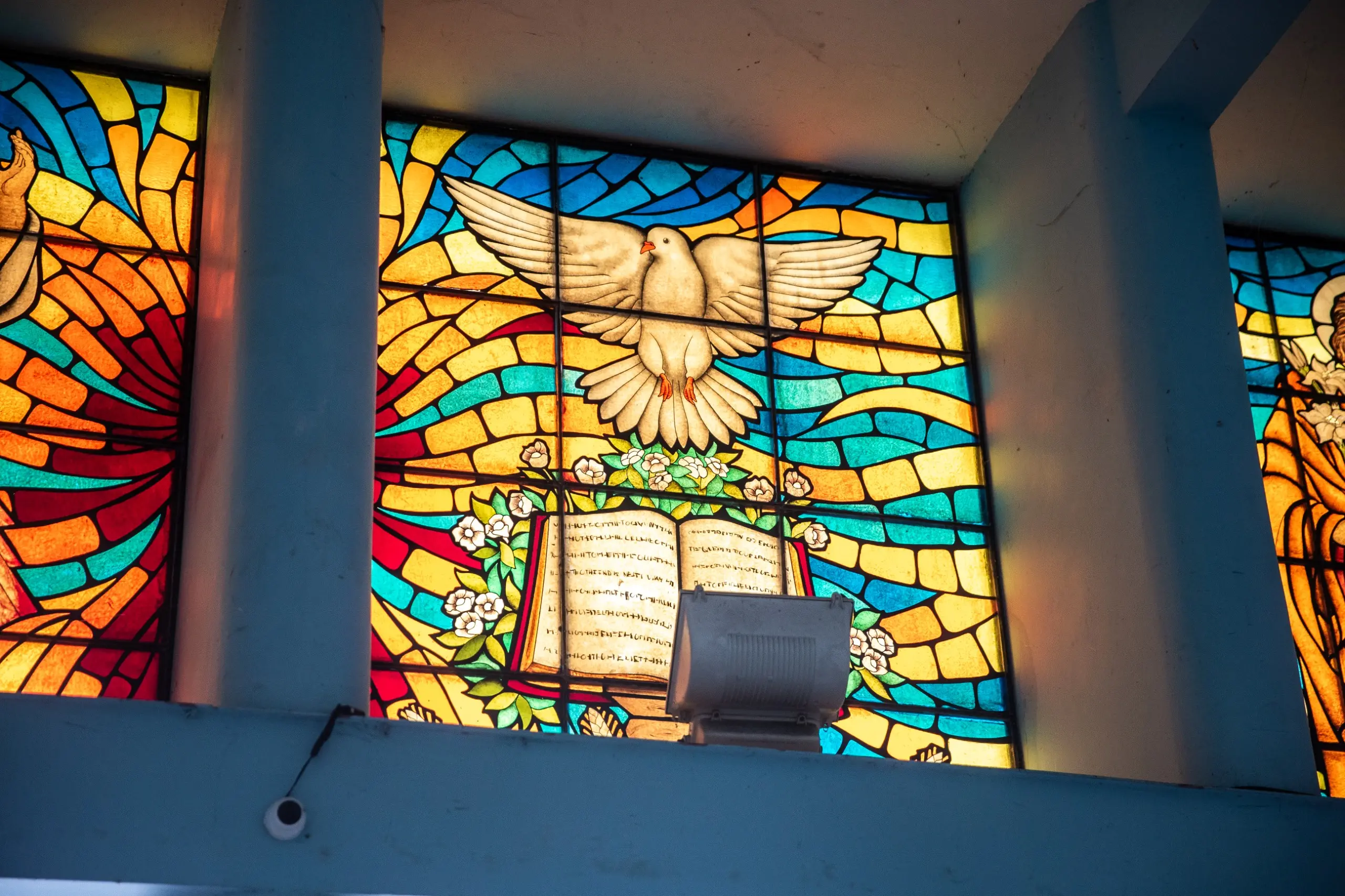 Uma fotografia de um vitral menor dentro da igreja. O vitral mostra uma pomba branca, representando o Espírito Santo, voando sobre um livro aberto decorado com flores. O fundo é uma composição de formas geométricas em tons de azul, amarelo, vermelho e turquesa
