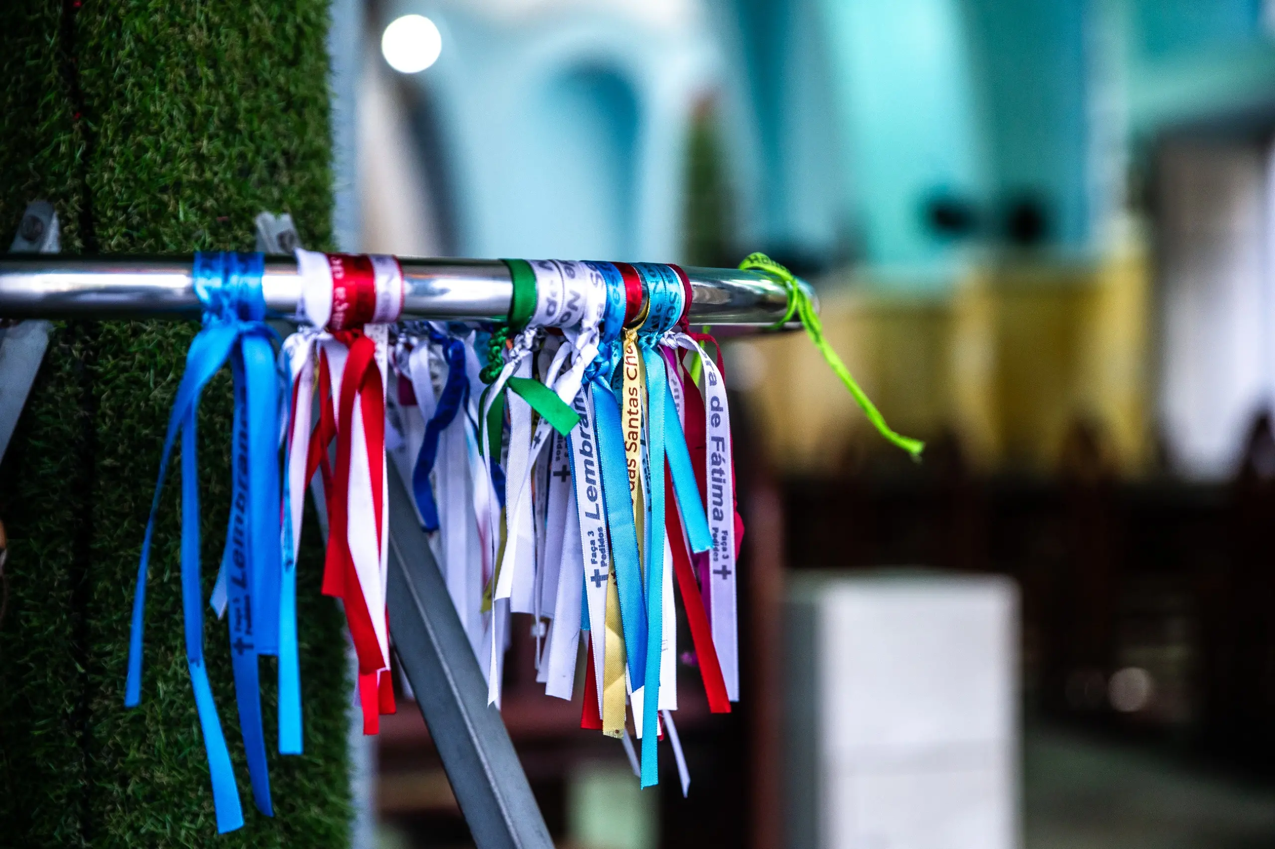 Uma fotografia de fitas coloridas, semelhantes às fitas do Senhor do Bonfim, amarradas em um corrimão de metal. As fitas têm diferentes cores e algumas delas têm inscrições, como 