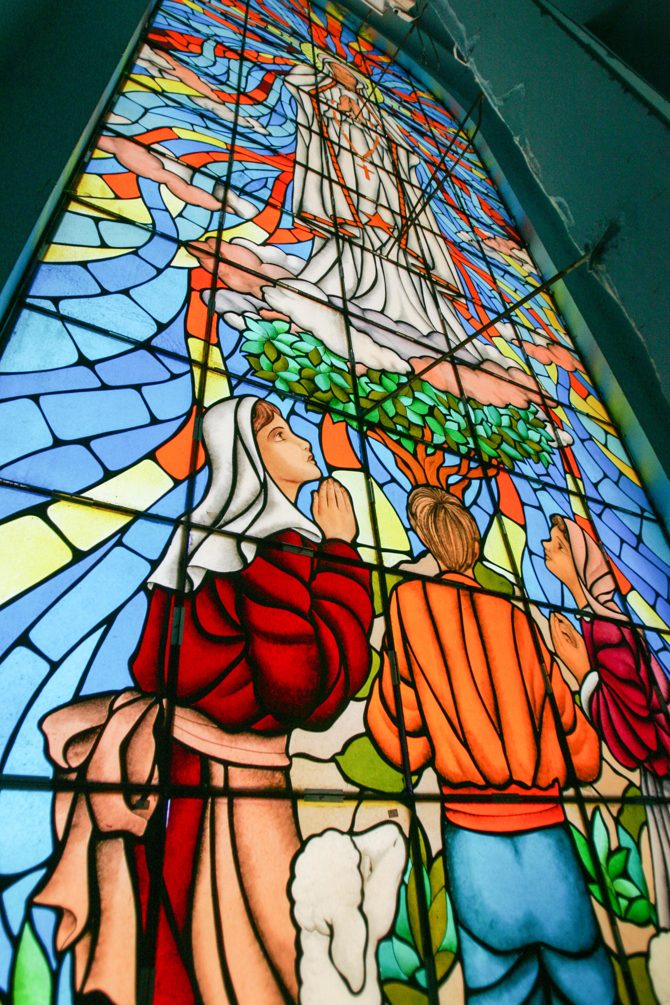 Uma fotografia de um grande vitral colorido dentro da Igreja de Fátima. O vitral retrata a aparição de Nossa Senhora de Fátima, com a Virgem Maria em vestes brancas e luminosas no topo, e as três crianças, Lúcia, Francisco e Jacinta, em oração, olhando para cima. As cores do vitral são vivas, predominando o azul, vermelho, laranja e amarelo