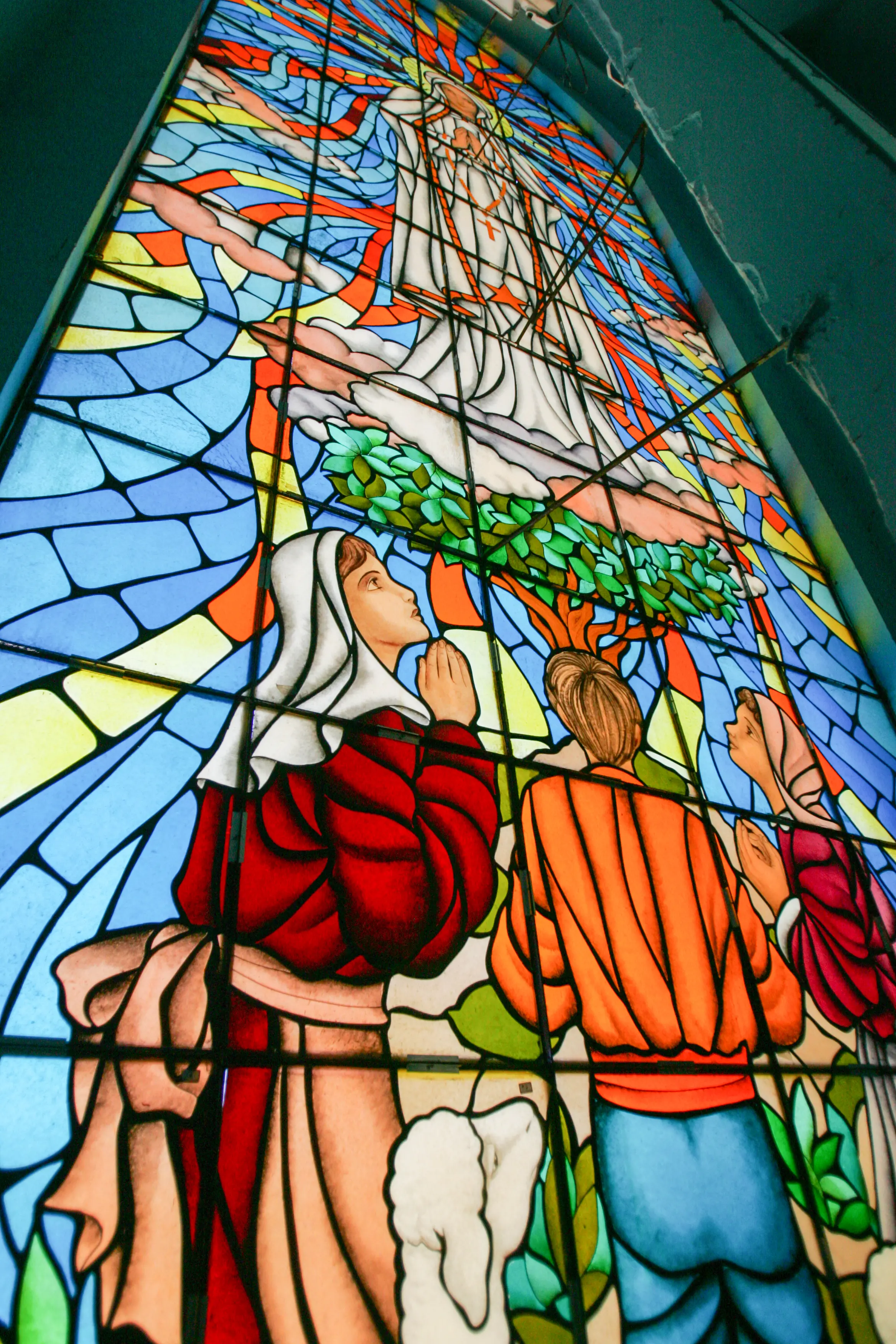 Uma fotografia de um grande vitral colorido dentro da Igreja de Fátima. O vitral retrata a aparição de Nossa Senhora de Fátima, com a Virgem Maria em vestes brancas e luminosas no topo, e as três crianças, Lúcia, Francisco e Jacinta, em oração, olhando para cima. As cores do vitral são vivas, predominando o azul, vermelho, laranja e amarelo