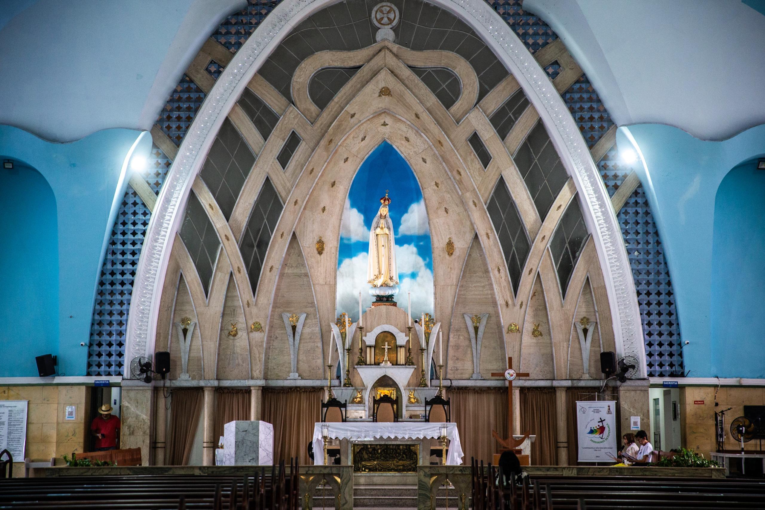 Uma fotografia do interior da Igreja de Fátima, mostrando o altar principal de frente. A arquitetura interna é moderna, com arcos e linhas que se encontram no teto. Uma grande estátua de Nossa Senhora de Fátima está em um nicho azul com nuvens, no centro do altar. O altar principal, com uma toalha branca, está em frente às cadeiras do clero. Bancos de madeira para os fiéis estão em primeiro plano