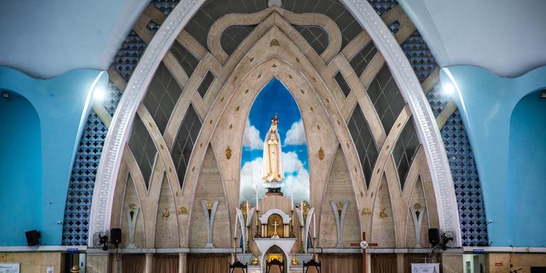 Uma fotografia do interior da Igreja de Fátima, mostrando o altar principal de frente. A arquitetura interna é moderna, com arcos e linhas que se encontram no teto. Uma grande estátua de Nossa Senhora de Fátima está em um nicho azul com nuvens, no centro do altar. O altar principal, com uma toalha branca, está em frente às cadeiras do clero. Bancos de madeira para os fiéis estão em primeiro plano