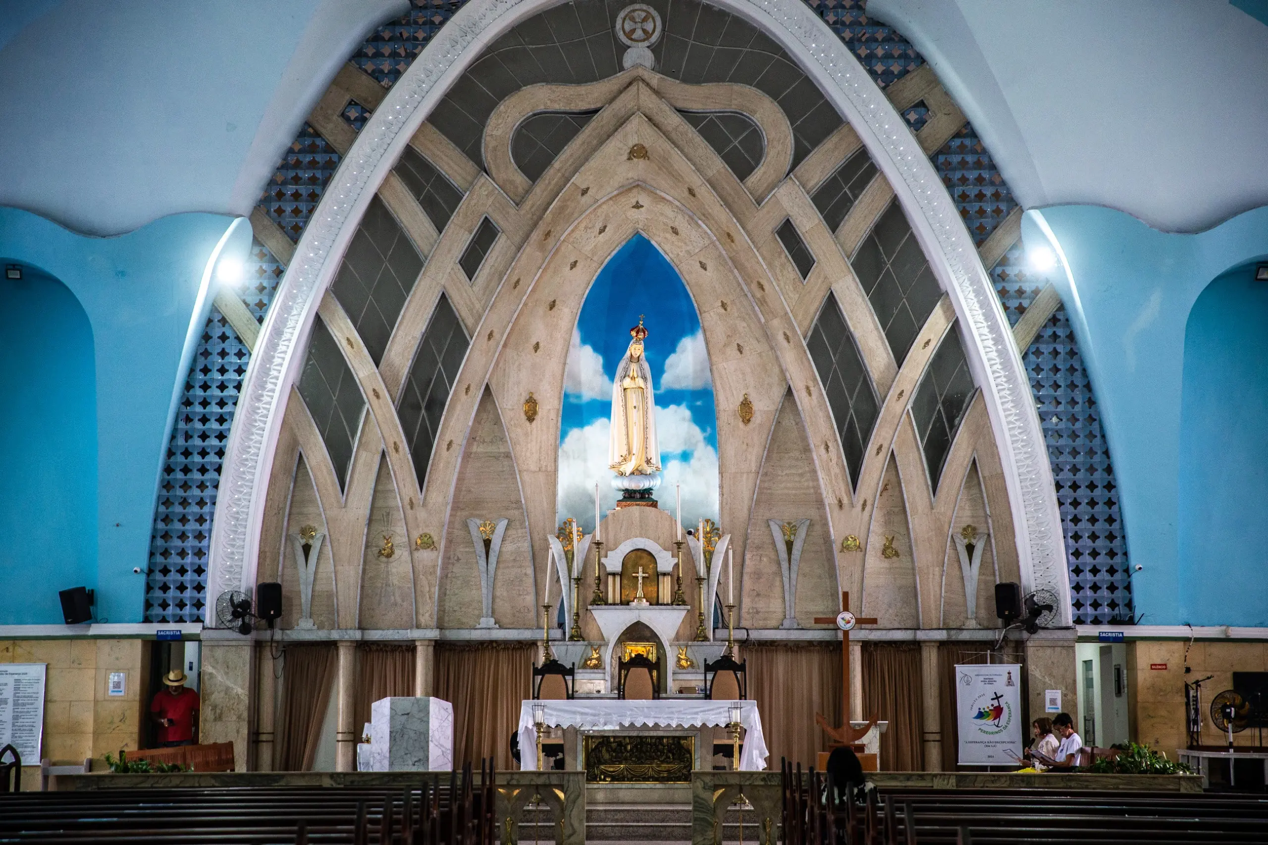 Uma fotografia do interior da Igreja de Fátima, mostrando o altar principal de frente. A arquitetura interna é moderna, com arcos e linhas que se encontram no teto. Uma grande estátua de Nossa Senhora de Fátima está em um nicho azul com nuvens, no centro do altar. O altar principal, com uma toalha branca, está em frente às cadeiras do clero. Bancos de madeira para os fiéis estão em primeiro plano