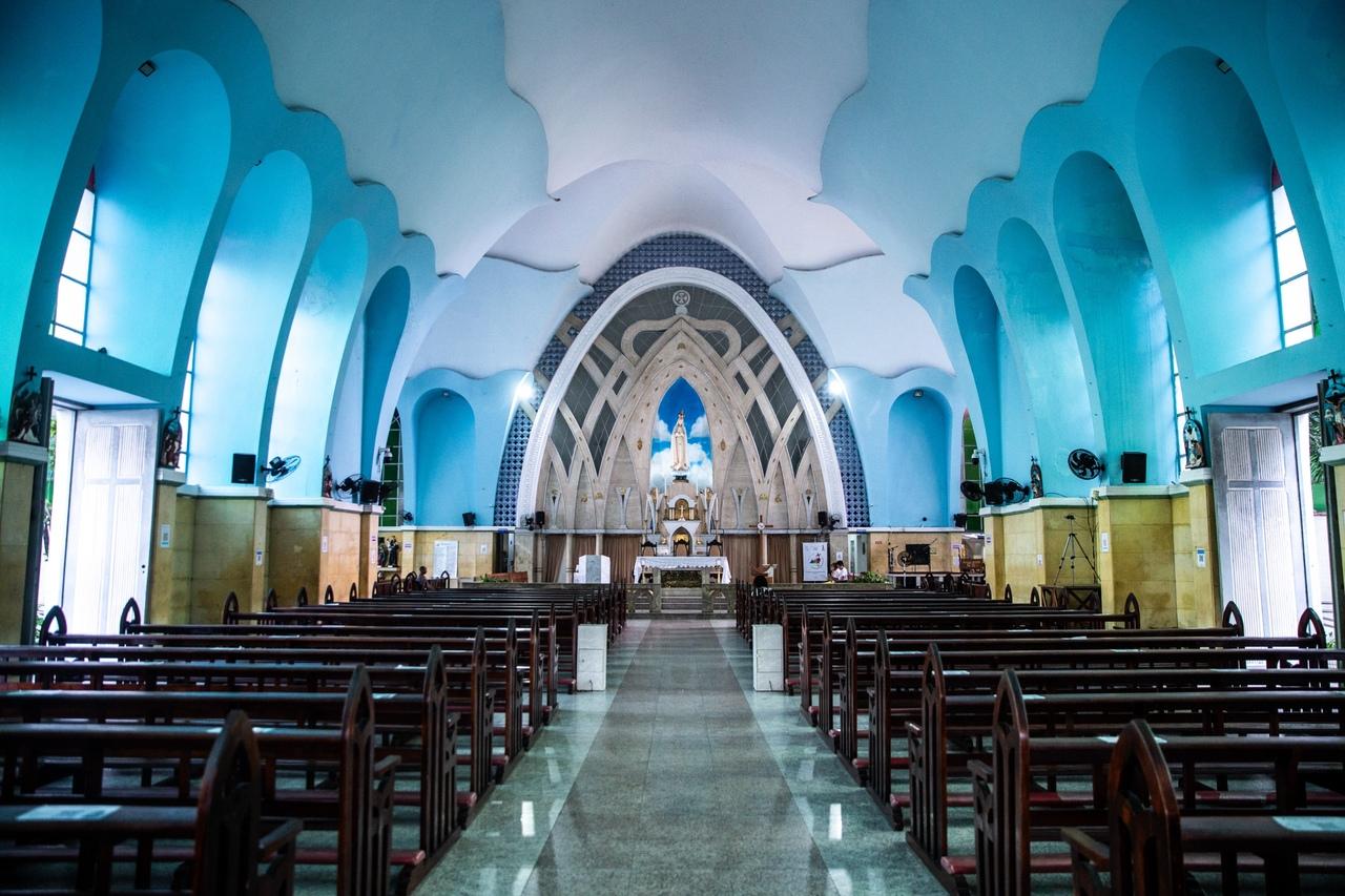 Uma fotografia em preto e branco mostra a Igreja de Fátima em Fortaleza. A igreja tem uma arquitetura moderna com arcos curvados na fachada e uma torre sineira simples com uma cruz no topo. Uma grande rosário pendurado da torre é visível. O céu está nublado, e o primeiro plano é de um terreno com vegetação esparsa. Na parte inferior da foto, há uma legenda manuscrita que diz 