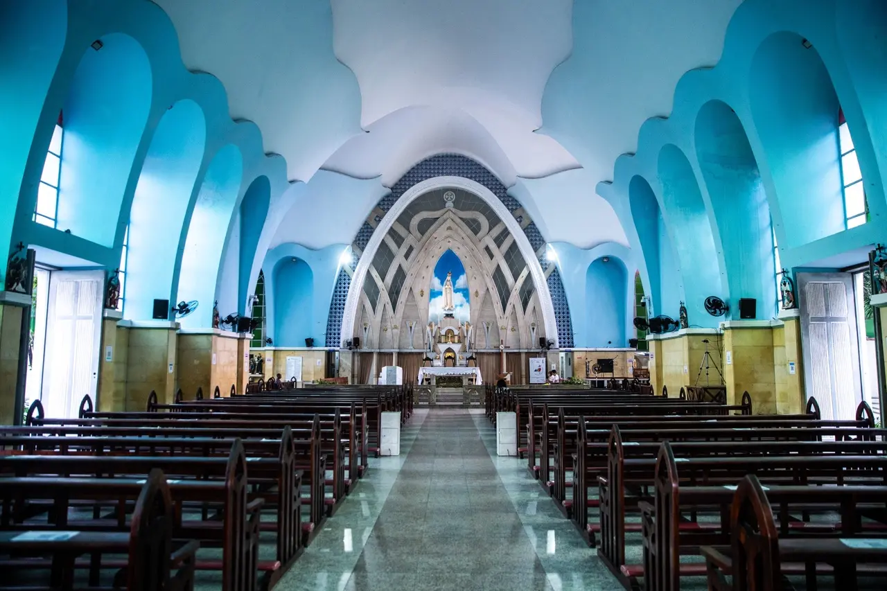 Uma fotografia em preto e branco mostra a Igreja de Fátima em Fortaleza. A igreja tem uma arquitetura moderna com arcos curvados na fachada e uma torre sineira simples com uma cruz no topo. Uma grande rosário pendurado da torre é visível. O céu está nublado, e o primeiro plano é de um terreno com vegetação esparsa. Na parte inferior da foto, há uma legenda manuscrita que diz 