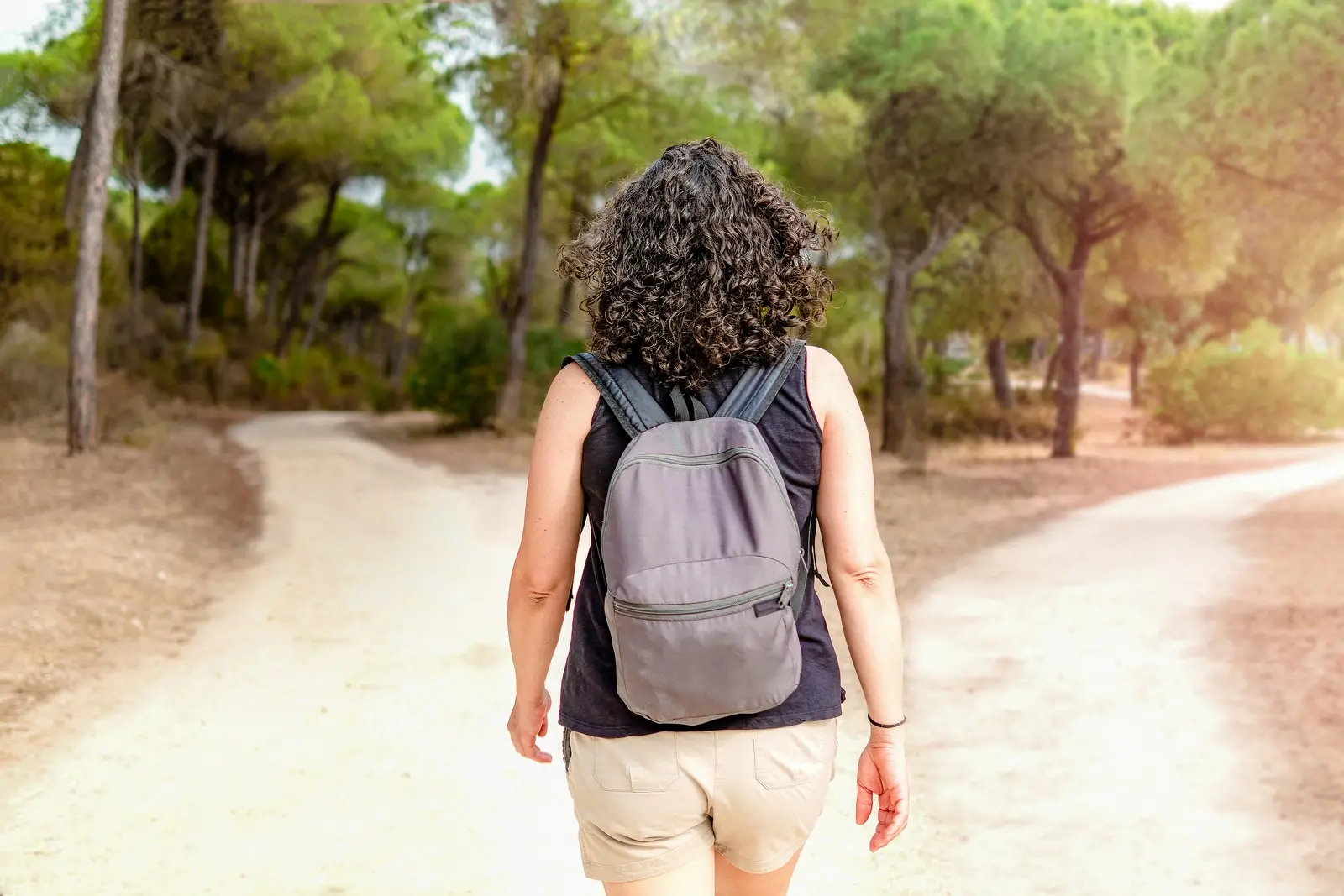 Imagem mostra mulher de costas, com mochila nas costas, short bege e blusa preta. Ela é branca, tem os cabelos curtos cacheados, e está diante de uma bifurcação entre dois caminhos. Ao fundo, uma floresta bem verde aparece.