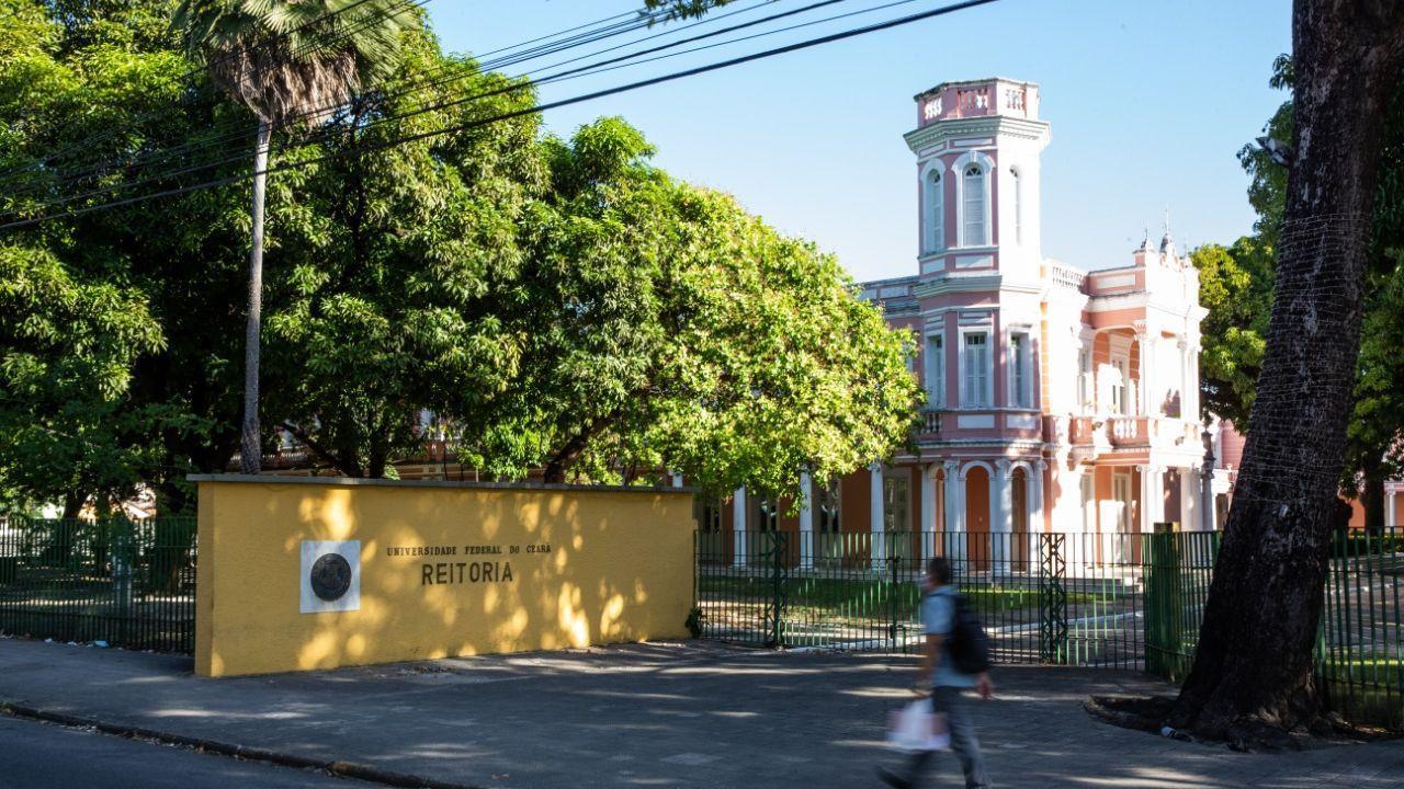 Imagem mostra fachada da reitoria da Universidade Federal do Ceará, no bairro Benfica, em Fortaleza. Atrás de um muro baixo amarelo e de grades verdes, está o prédio rosa com branco em arquitetura antiga. Um pedestre passa na calçada.