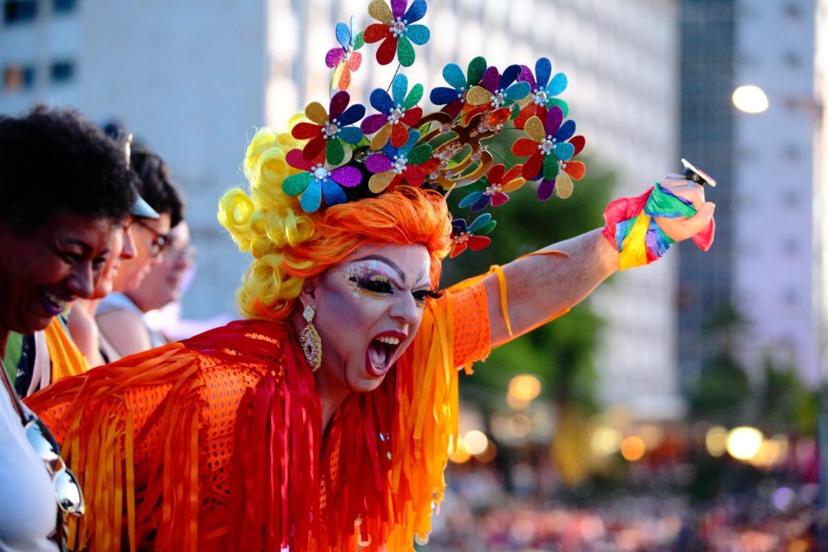 Pessoa vestida com fantasia vibrante nas cores do arco-íris, maquiagem artística e adorno de cabeça com flores coloridas, participa de um evento LGBTI+ em espaço urbano, rodeada por outras pessoas sorridentes