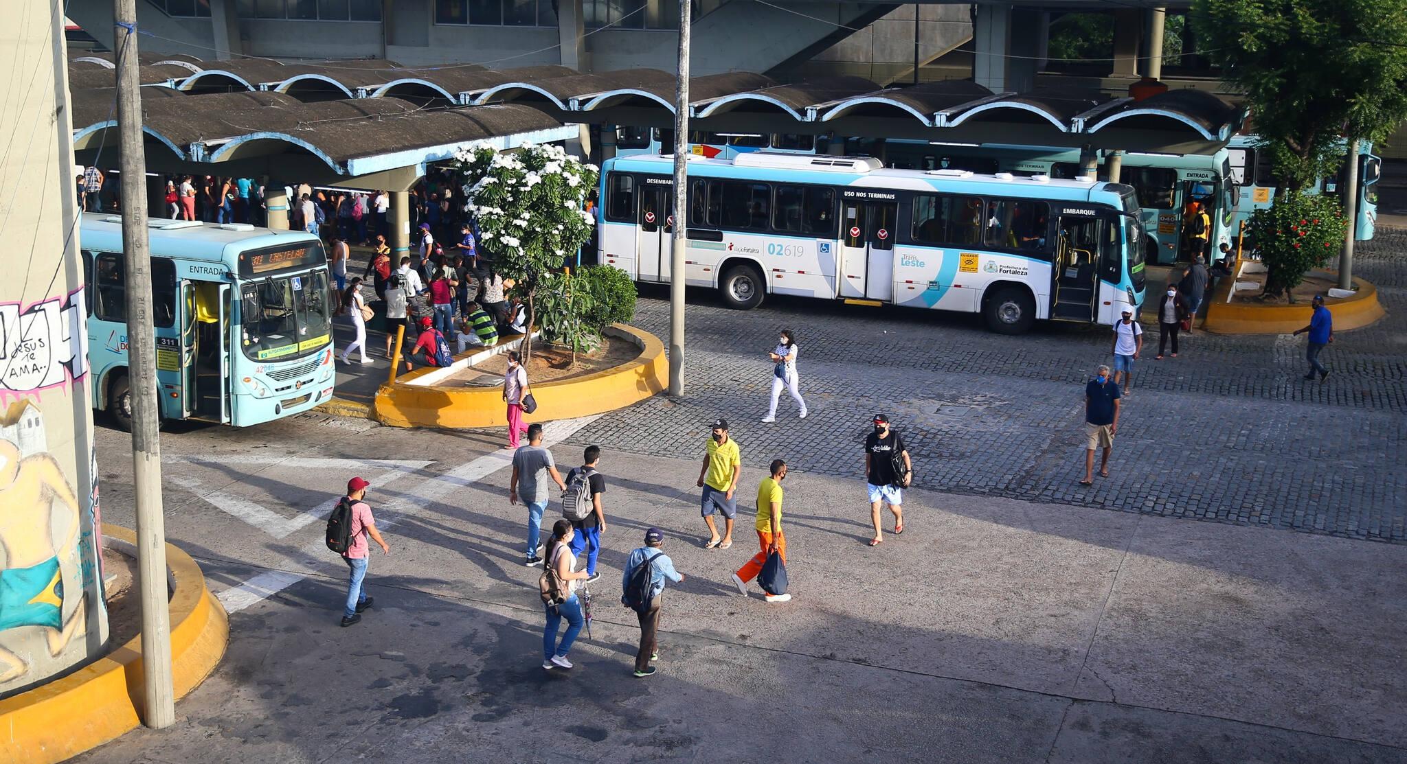 A imagem mostra um terminal de ônibus movimentado, com pessoas caminhando em uma área aberta. Dois ônibus estão parados nas plataformas, onde há telhados arqueados. Ao fundo, um prédio de concreto e uma estrutura de metal compõem o ambiente urbano.
