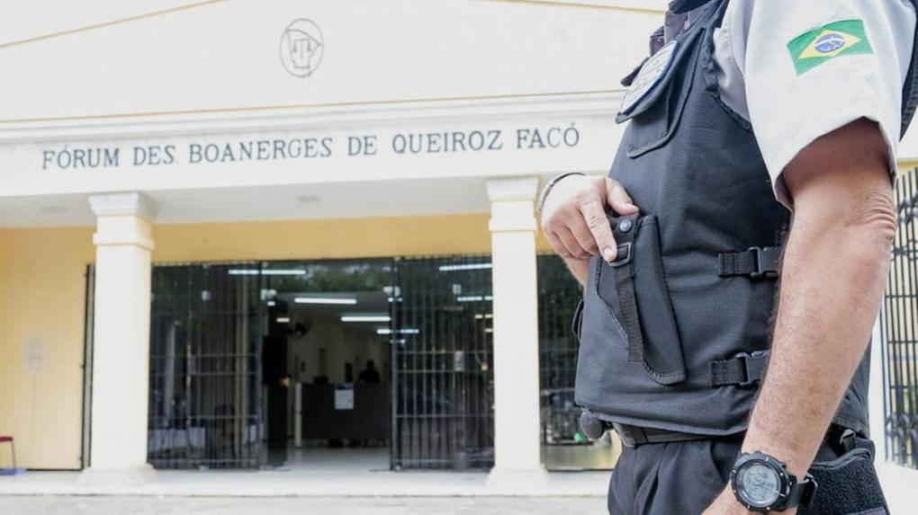 Imagem mostra o Forum Desembargador Boanerges de Queiroz Facó ao fundo, em Iguatu, e um agente de segurança em primeiro plano, com colete e segurando uma arma de fogo
