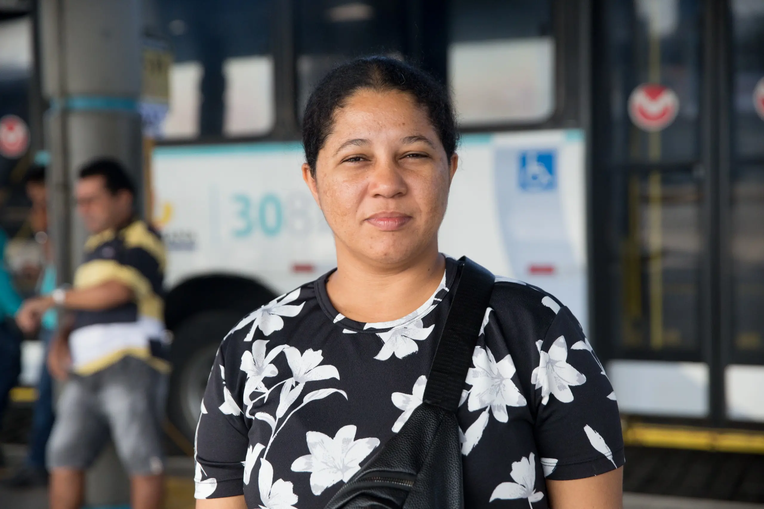 Imagem mostra mulher de cabelo curto em primeiro plano, vestindo blusa preta com flores brancas. Ao fundo, um ônibus estacionado.