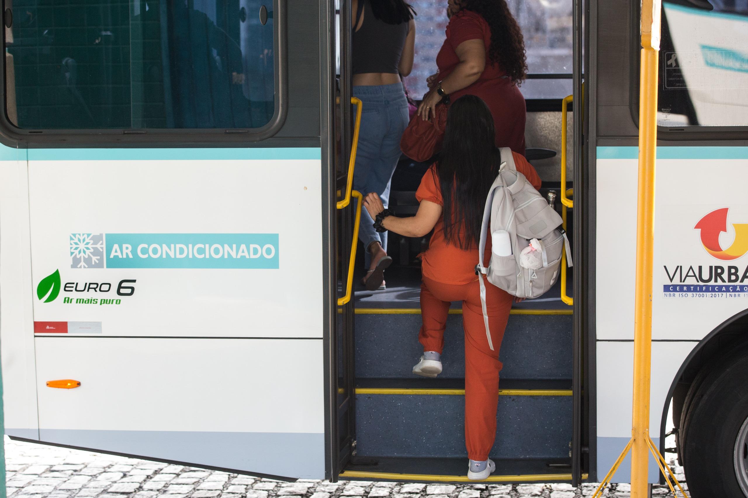 Imagem mostra mulher subindo degraus de ônibus. Ela está de costas e veste calça laranja e blusa preta.