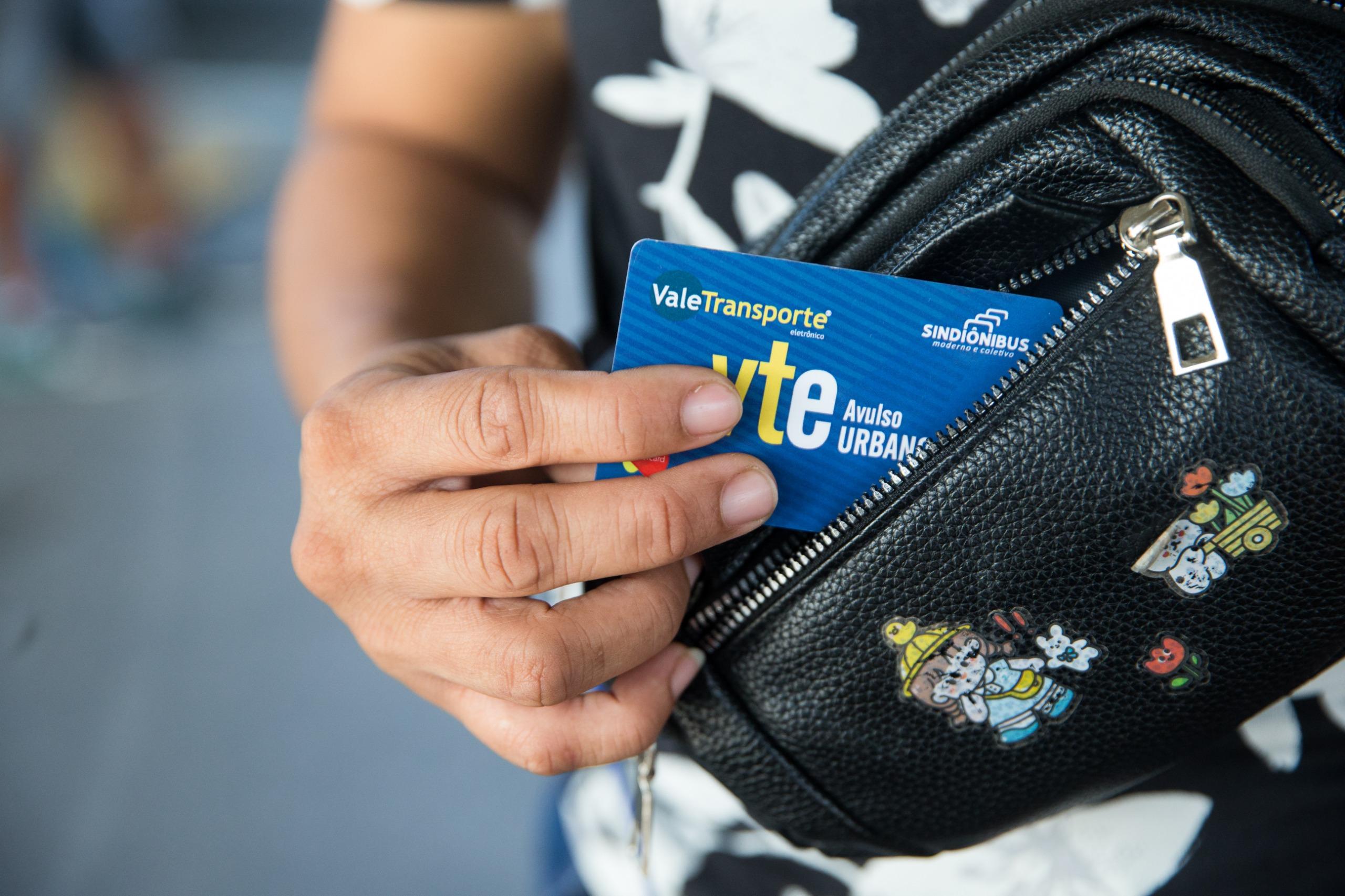 Imagem mostra mão tirando cartão de ônibus azuk de dentro de uma bolsa preta, em close.