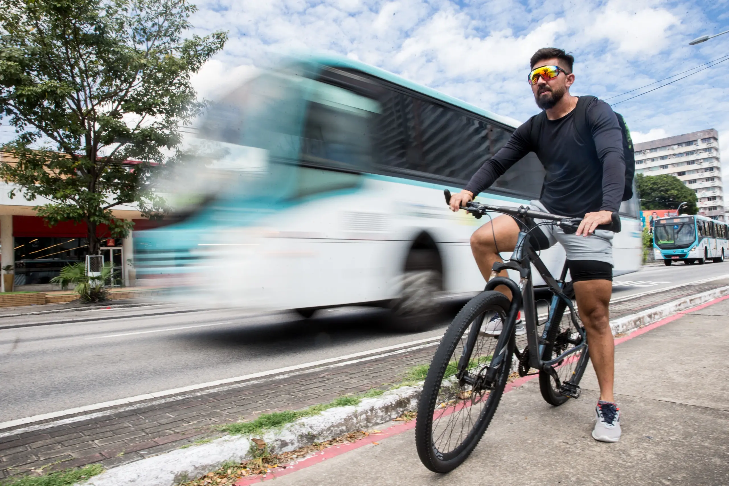 Imagem mostra homem sobre bicicleta. Ele veste short cinza, camisa de manga longa preta e tênis. Ao fundo, um ônibus embaçado em movimento.