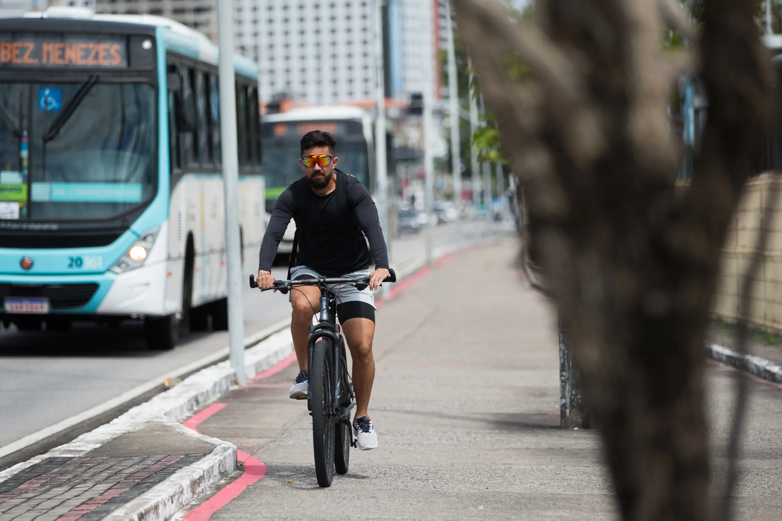 Imagem mostra homem sobre bicicleta. Ele veste short cinza, camisa de manga longa preta e tênis. Ao lado, um ônibus trafega na avenida.