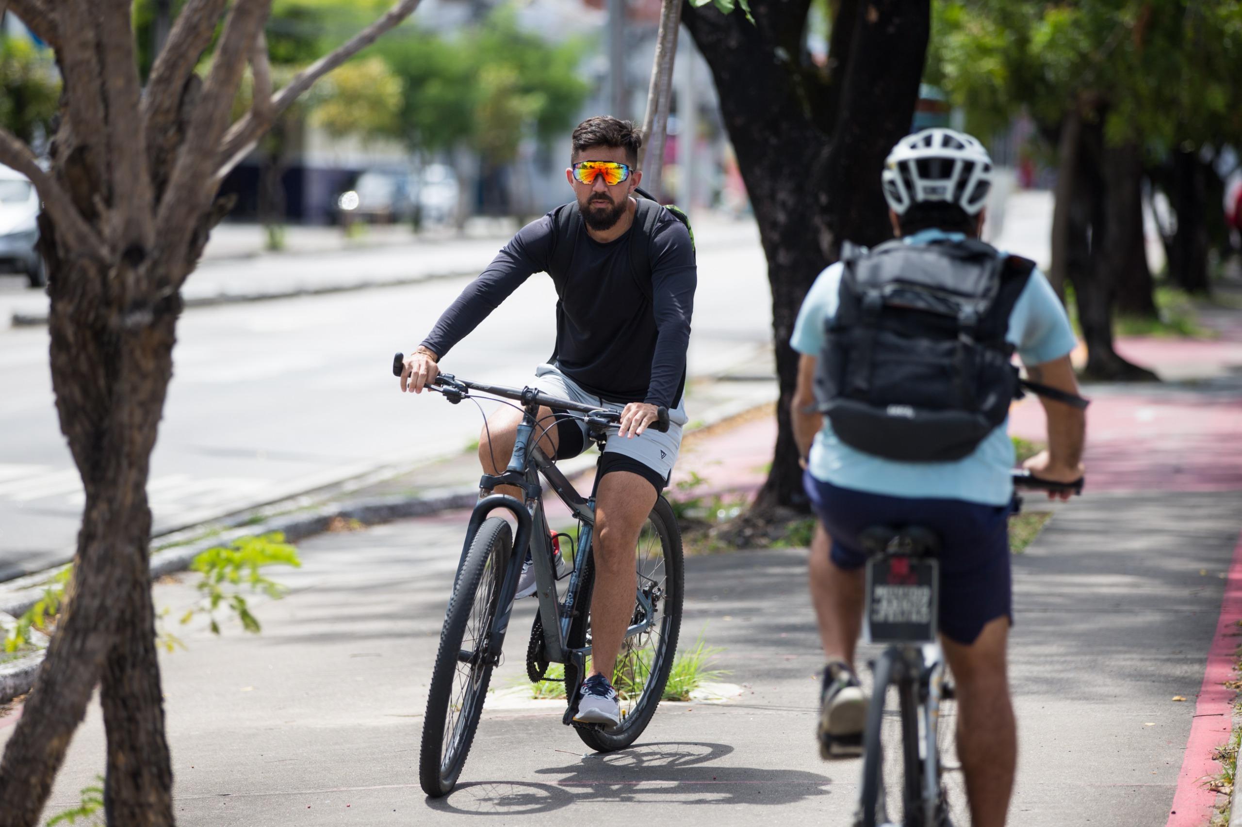 Imagem mostra homem sobre bicicleta. Ele veste short cinza, camisa de manga longa preta e tênis. De costas, outro homem de bicicleta e mochila nas costas.