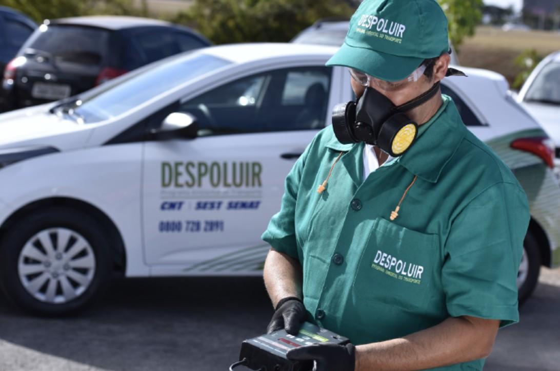 Imagem mostra um técnico usando um uniforme verde do programa DESPOLUIR, um boné e uma máscara de proteção com filtros amarelos, luvas pretas e óculos. Ele está segurando um equipamento eletrônico de medição em suas mãos. Ao fundo, um carro branco do programa é visível.
