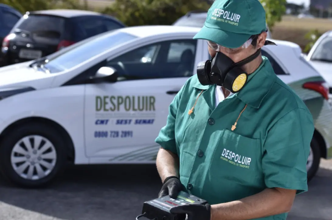 Imagem mostra um técnico usando um uniforme verde do programa DESPOLUIR, um boné e uma máscara de proteção com filtros amarelos, luvas pretas e óculos. Ele está segurando um equipamento eletrônico de medição em suas mãos. Ao fundo, um carro branco do programa é visível.