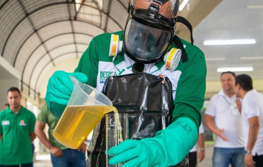 Imagem mostra uma pessoa usando um equipamento de proteção individual, incluindo um macacão verde e branco, máscara facial e luvas azuis. Ela derrama combustível de um grande copo medidor para um recipiente cilíndrico, para teste de qualidade.