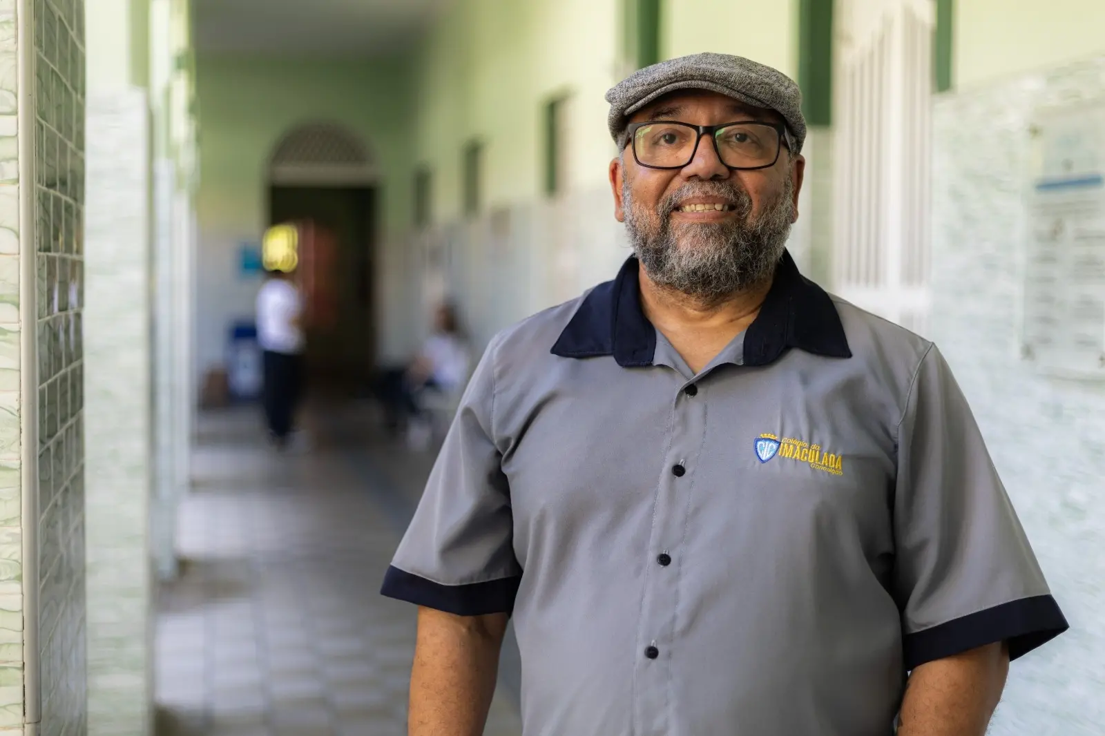 A imagem mostra um homem sorrindo, de barba grisalha e óculos, usando um boné de tecido cinza e um uniforme de gola polo cinza com detalhes escuros. O uniforme tem o brasão IMACULADA no peito. Ele está em um corredor bem iluminado, com paredes em tons claros, do Colégio da Imaculada Conceição.